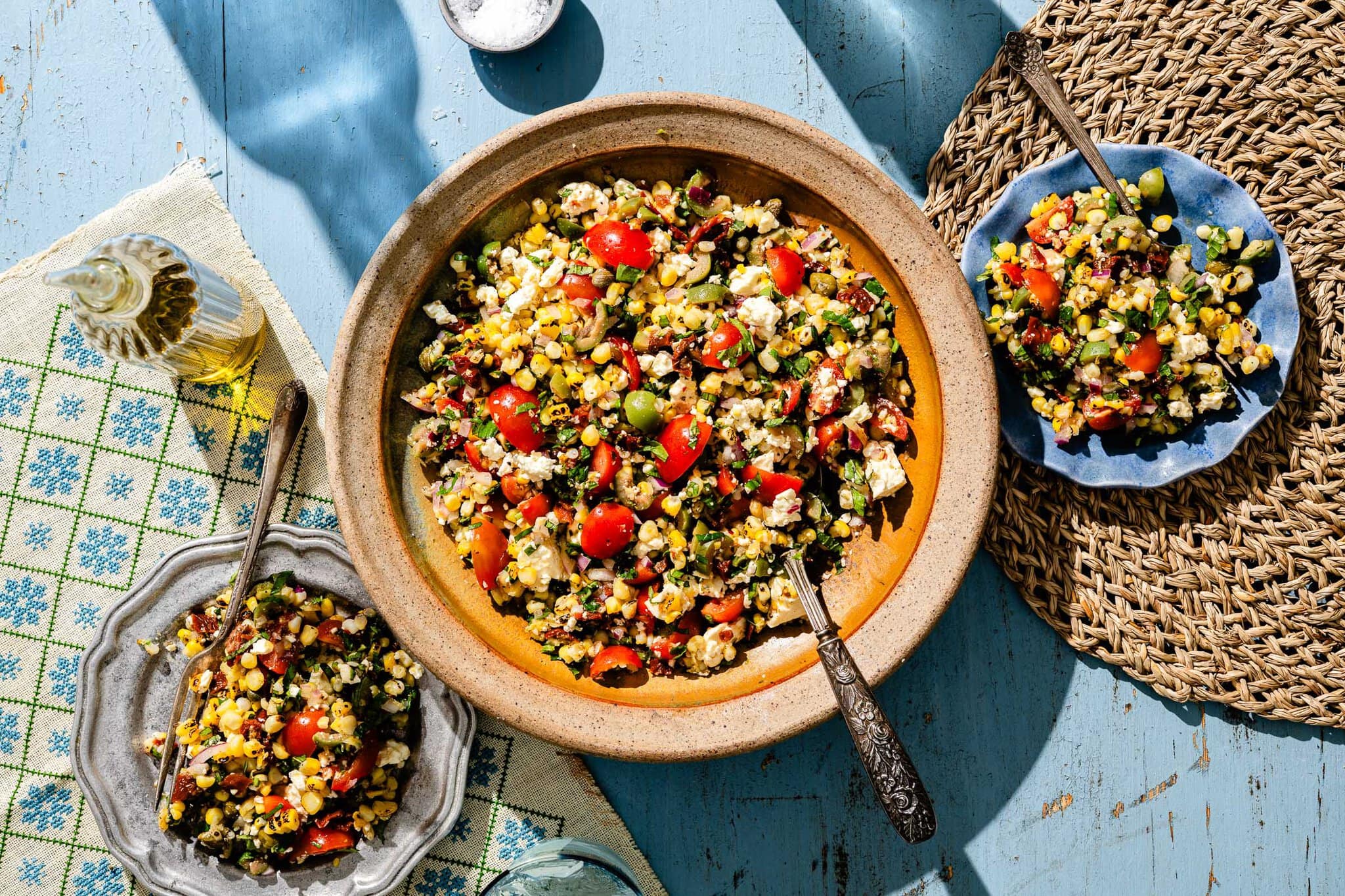 A large bowl and two smaller plates filled with a colorful salad containing corn, tomatoes, beans, green onions, and crumbled cheese, set on a blue wooden table with utensils and a bottle of olive oil nearby.