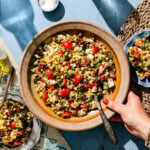 A hand holds a rustic bowl filled with a colorful salad of corn, tomatoes, feta, olives, and herbs on a blue table, with a plate of salad and salt nearby.