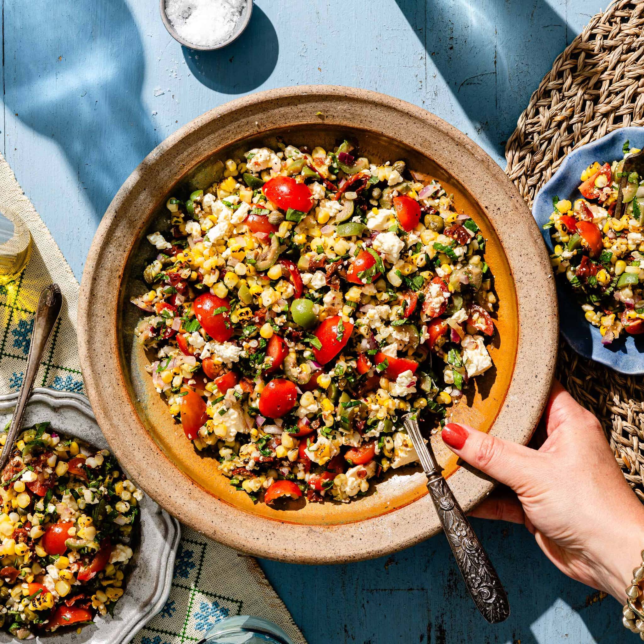 A hand holds a rustic bowl filled with a colorful salad of corn, tomatoes, feta, olives, and herbs on a blue table, with a plate of salad and salt nearby.