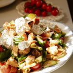 A plate of fresh salad with tomatoes, cucumbers, greens, croutons, and a dollop of creamy dressing, sprinkled with pepper. In the background, a plate of cherry tomatoes is visible.