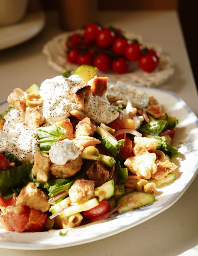A plate of fresh salad with tomatoes, cucumbers, greens, croutons, and a dollop of creamy dressing, sprinkled with pepper. In the background, a plate of cherry tomatoes is visible.