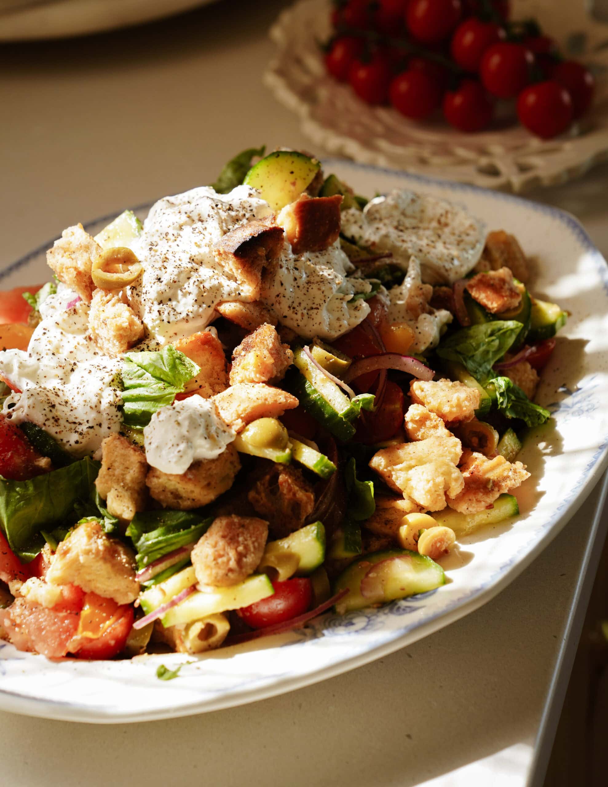 A salad with greens, tomatoes, cucumbers, olives, croutons, and dollops of creamy dressing, served on a white platter. A plate of cherry tomatoes is blurred in the background.