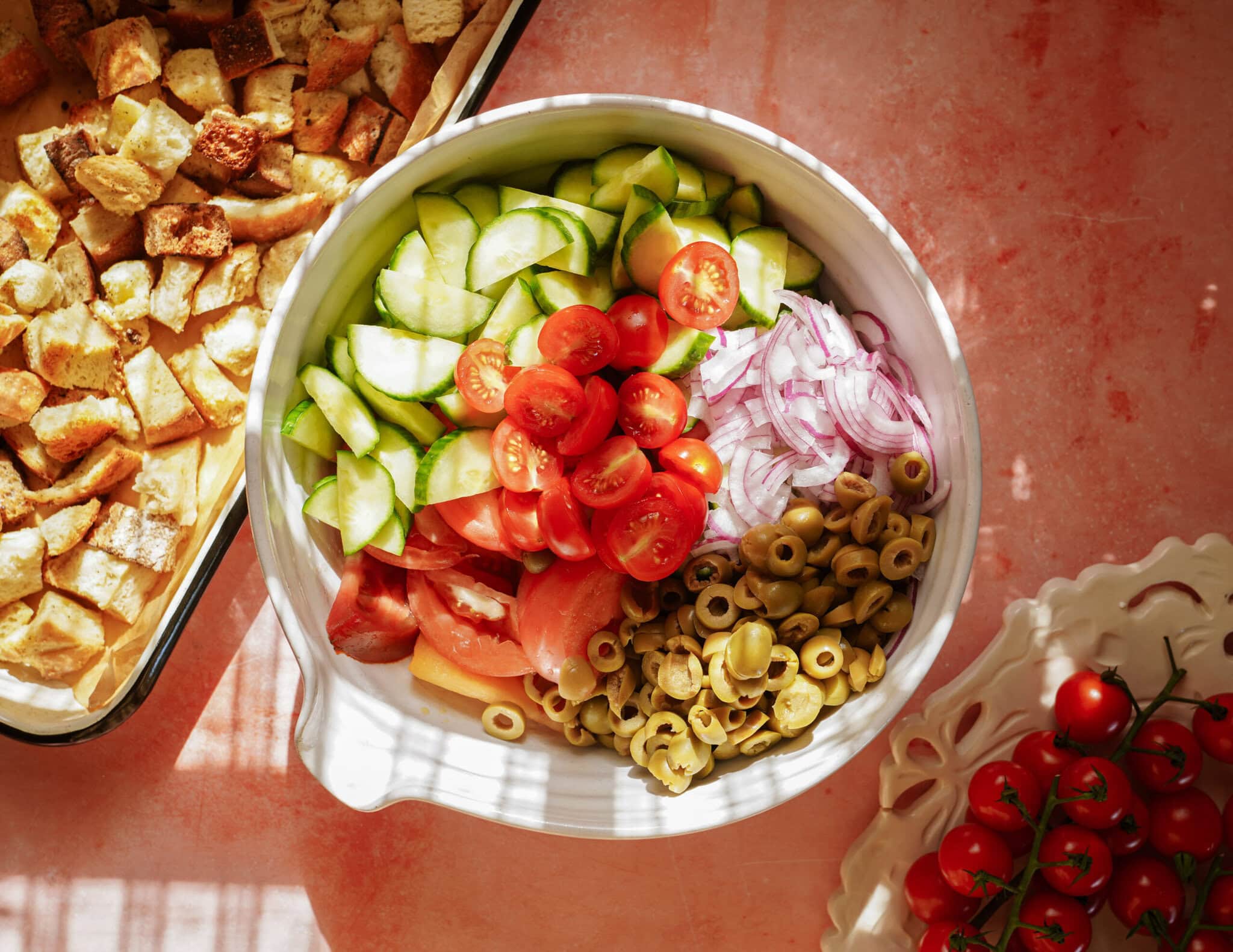 A bowl with sliced cucumbers, cherry tomatoes, red onions, green olives, and croutons, set on a pink surface next to more croutons in a tray and a small plate of cherry tomatoes.