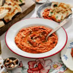 A bowl of red pepper dip garnished with chopped nuts, surrounded by pita bread slices on floral tablecloth with small plates and a bowl of nuts nearby.