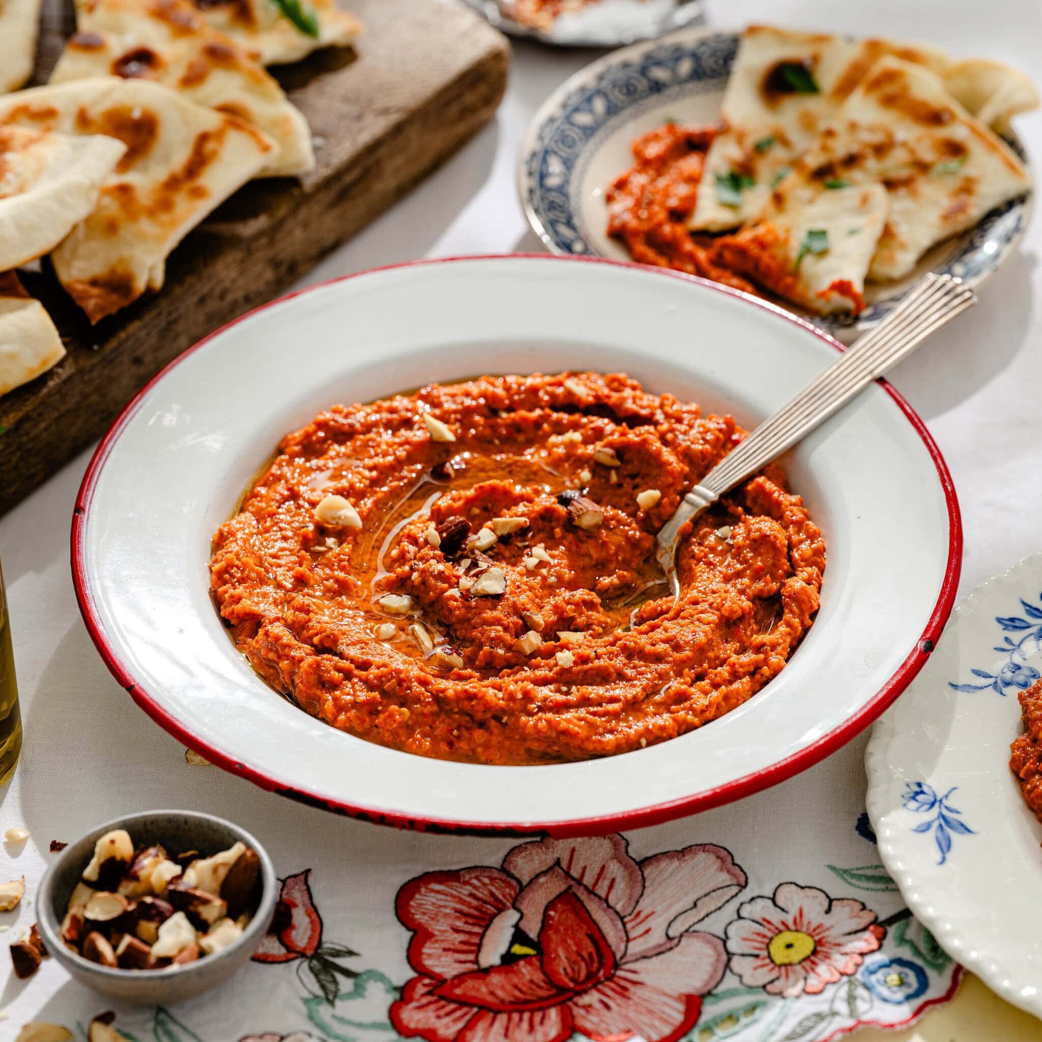 A bowl of red pepper dip garnished with chopped nuts, surrounded by pita bread slices on floral tablecloth with small plates and a bowl of nuts nearby.