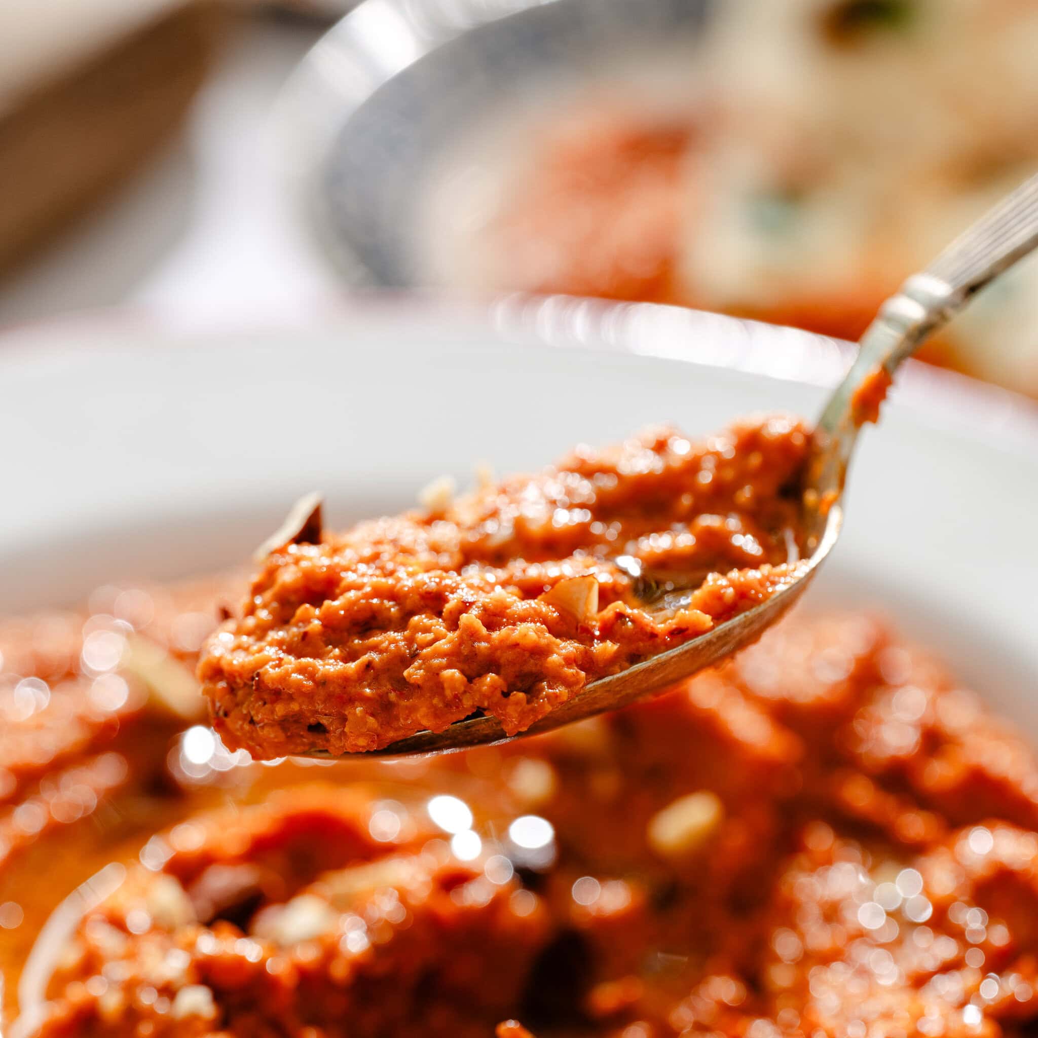 A close-up of a spoonful of thick, red-orange curry with visible spices and oil, held above a plate of the same curry in bright natural light.