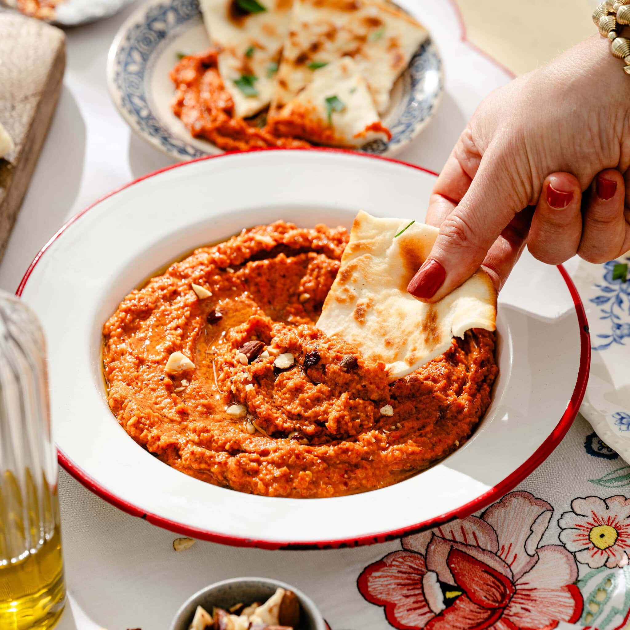 A hand with red nails dips naan bread into a bowl of red pepper dip, garnished with chopped nuts. Plates, more dip, and a glass bottle of olive oil are on a floral tablecloth in the background.