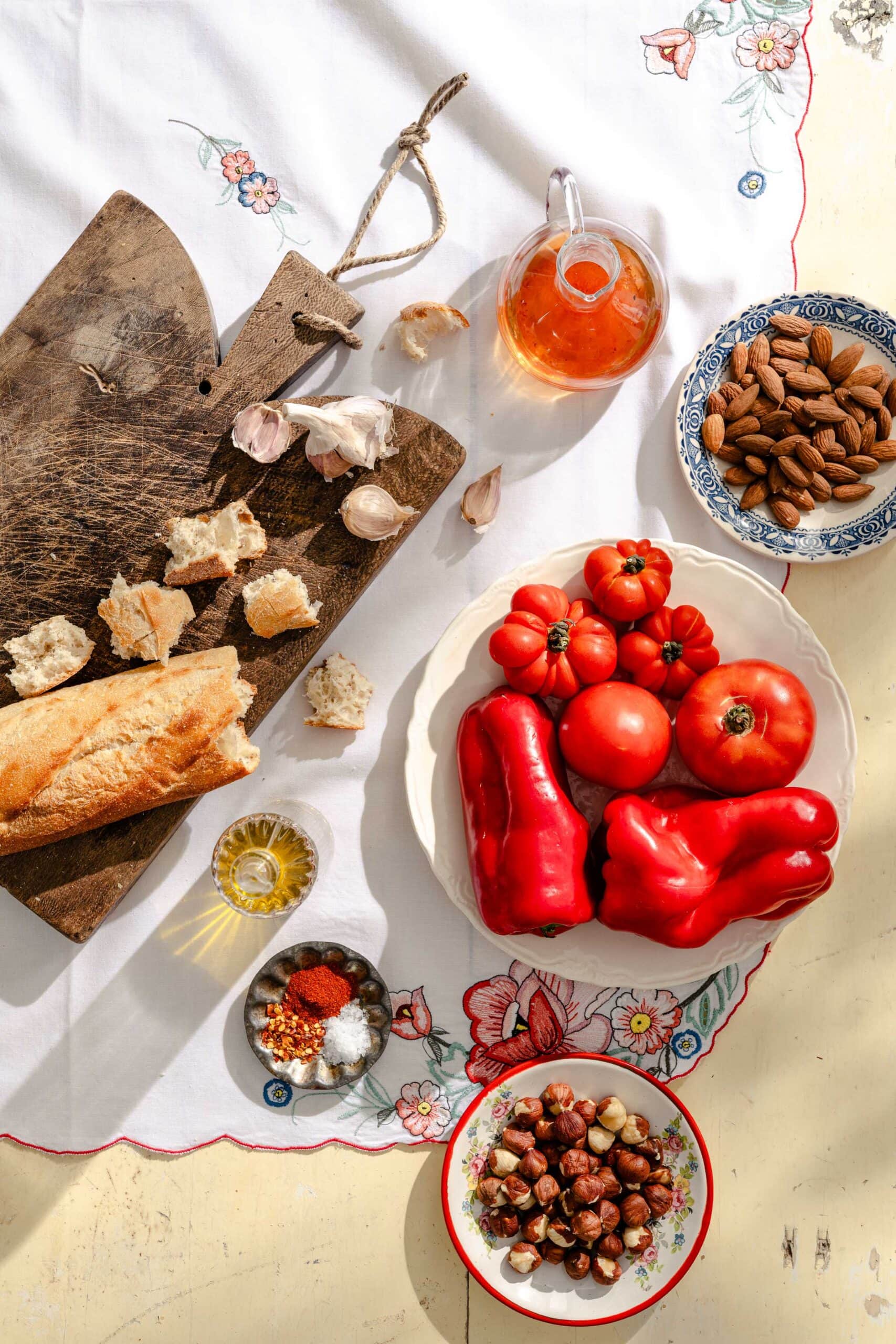 A top view of ingredients on a table: tomatoes, red bell peppers, baguette, nuts, almonds, spices, garlic, olive oil, and a glass jug of liquid, all on a floral tablecloth with a wooden cutting board.