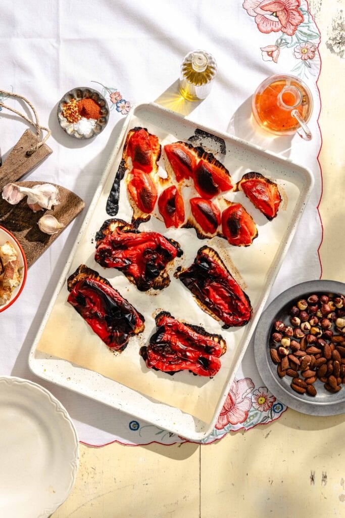 A tray of roasted red peppers on parchment paper, surrounded by garlic, spices, olive oil, nuts in a bowl, and a teapot, all arranged on a table with a floral tablecloth.