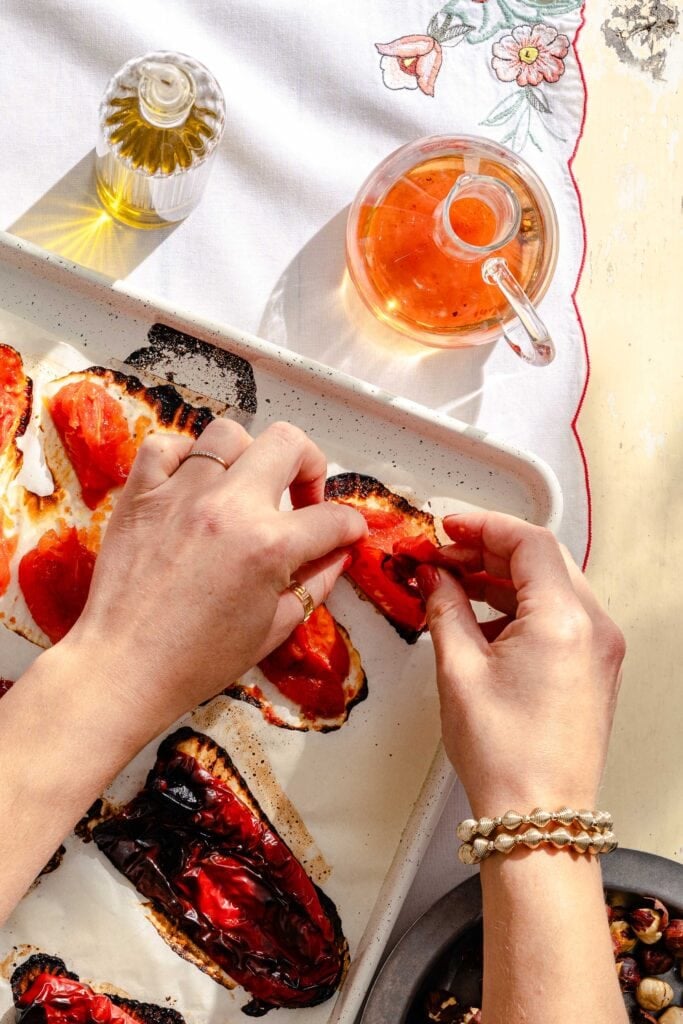 Hands preparing charred red peppers on toast, with a bottle of oil, a glass teapot with amber liquid, and a white cloth with floral embroidery on a sunlit table.