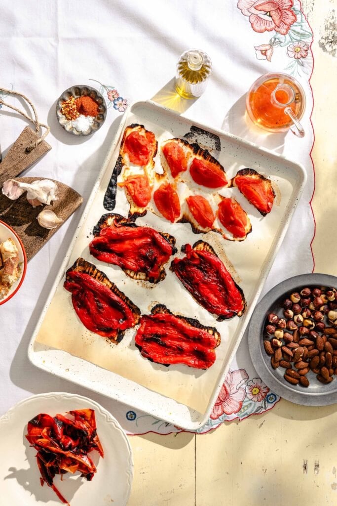 A baking tray with grilled eggplant slices topped with roasted red peppers and tomatoes, surrounded by small bowls of nuts, spices, garlic, olive oil, and a plate with more roasted peppers on a floral tablecloth.