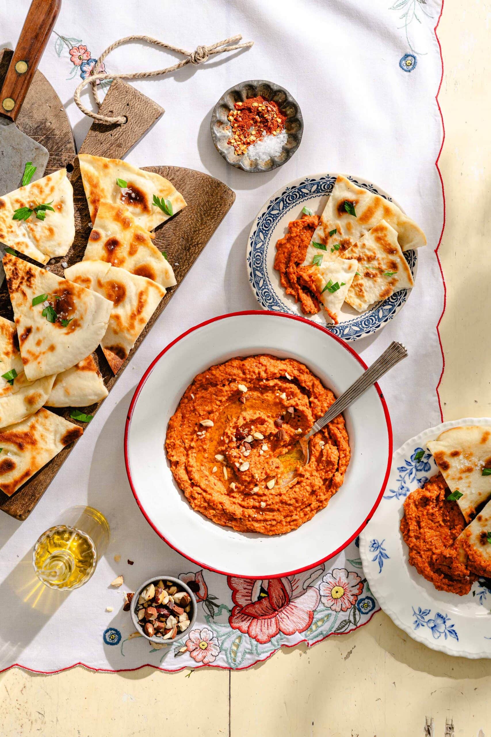 Bowl of red pepper hummus topped with crushed nuts, surrounded by pita bread slices on plates and a cutting board, with small bowls of spices and nuts on a floral tablecloth.