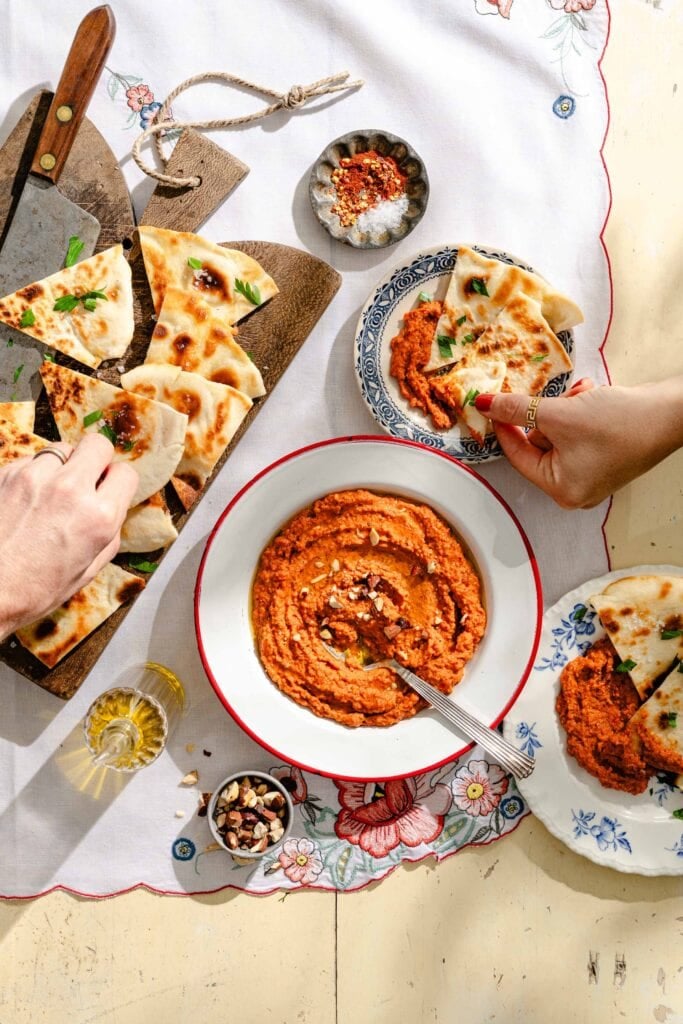 Two people dip pieces of flatbread into a bowl of red dip on a floral tablecloth, with extra flatbread, seasonings, and olive oil nearby.