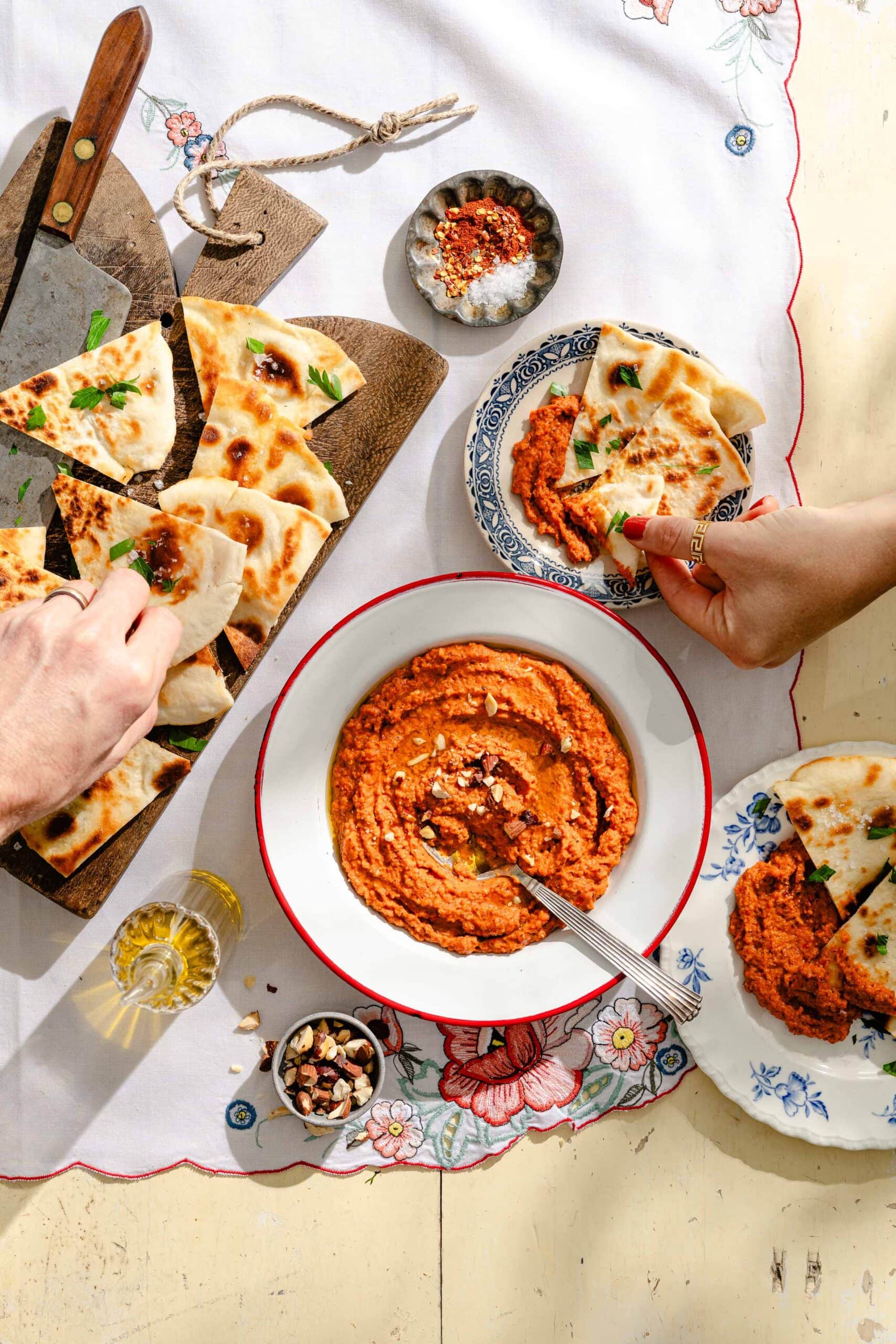 Two people dip pieces of flatbread into a bowl of red dip on a floral tablecloth, with extra flatbread, seasonings, and olive oil nearby.