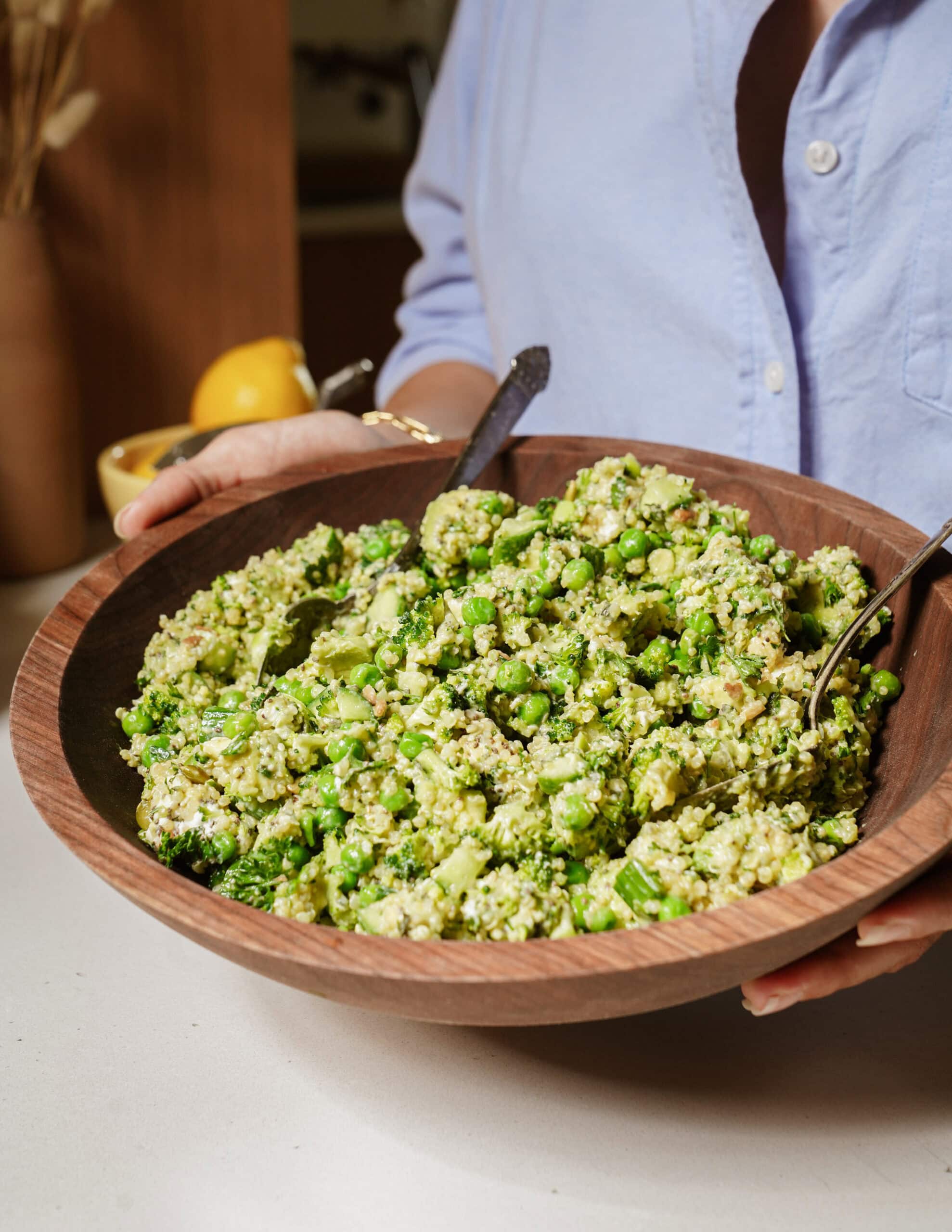 Person holding a large wooden bowl filled with quinoa, broccoli, and green vegetables, with two serving spoons. The person is wearing a light blue shirt, and the background shows a kitchen setting.