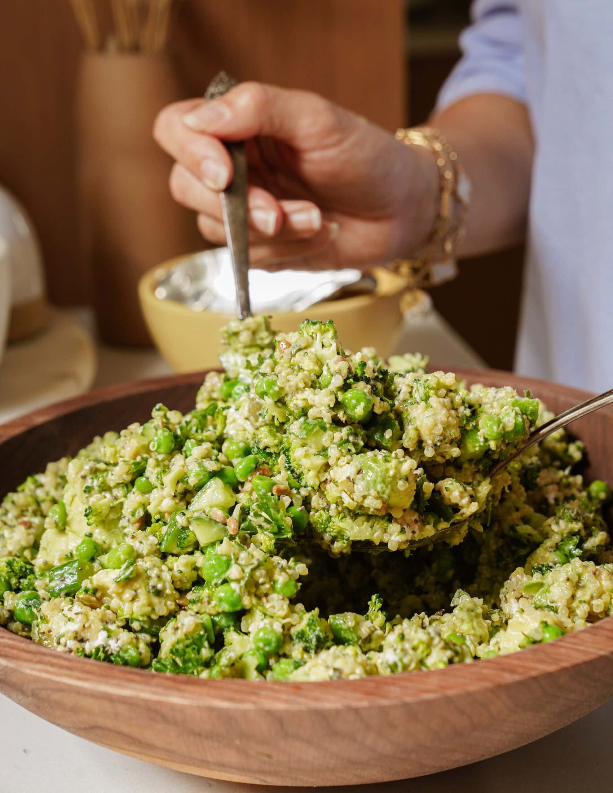 A person serves a green quinoa salad with broccoli and peas from a wooden bowl using two utensils. The salad looks fresh and healthy.