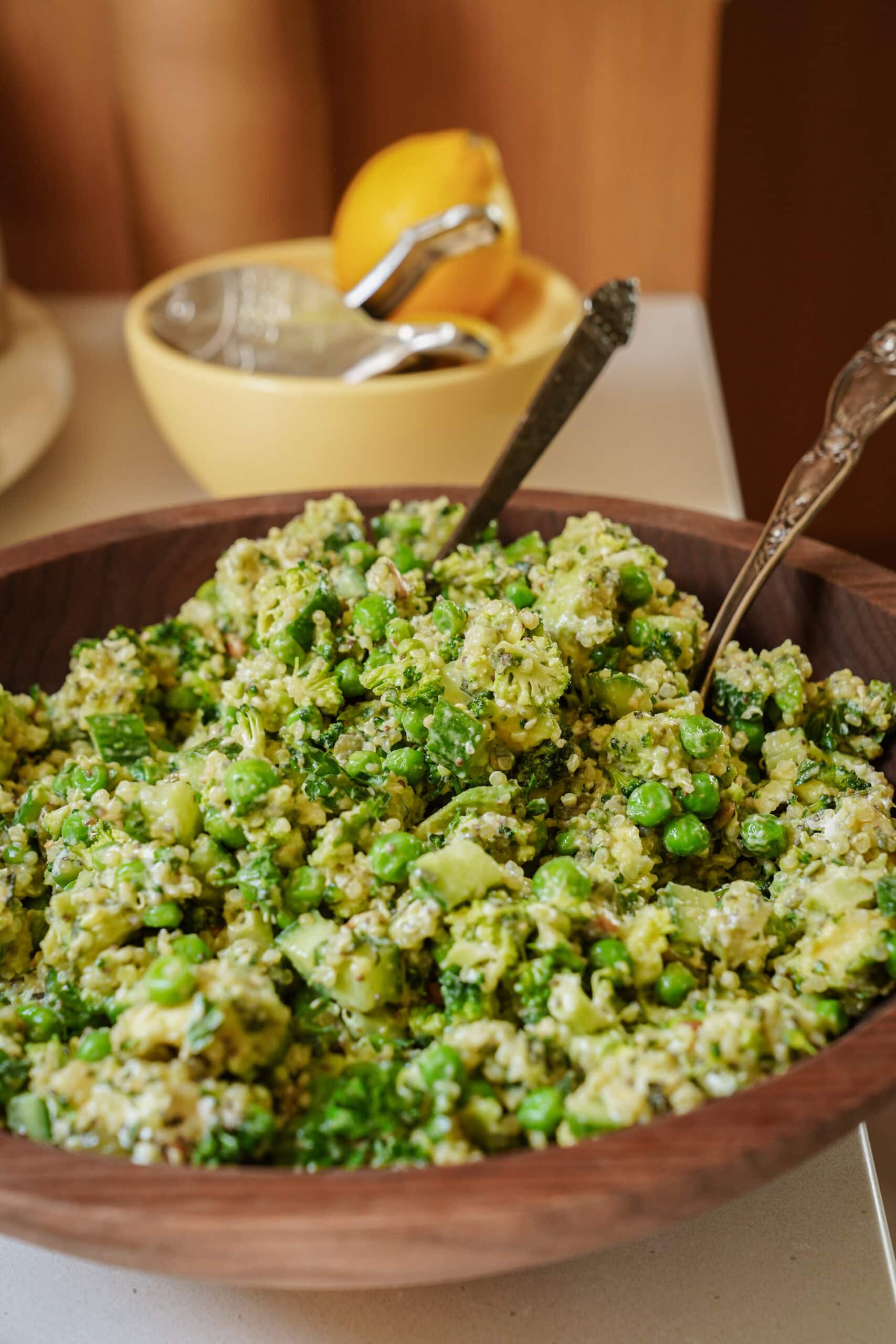 A wooden bowl filled with a green quinoa salad containing peas and chopped vegetables, with two serving utensils. A lemon juicer and a halved lemon are blurred in the background.