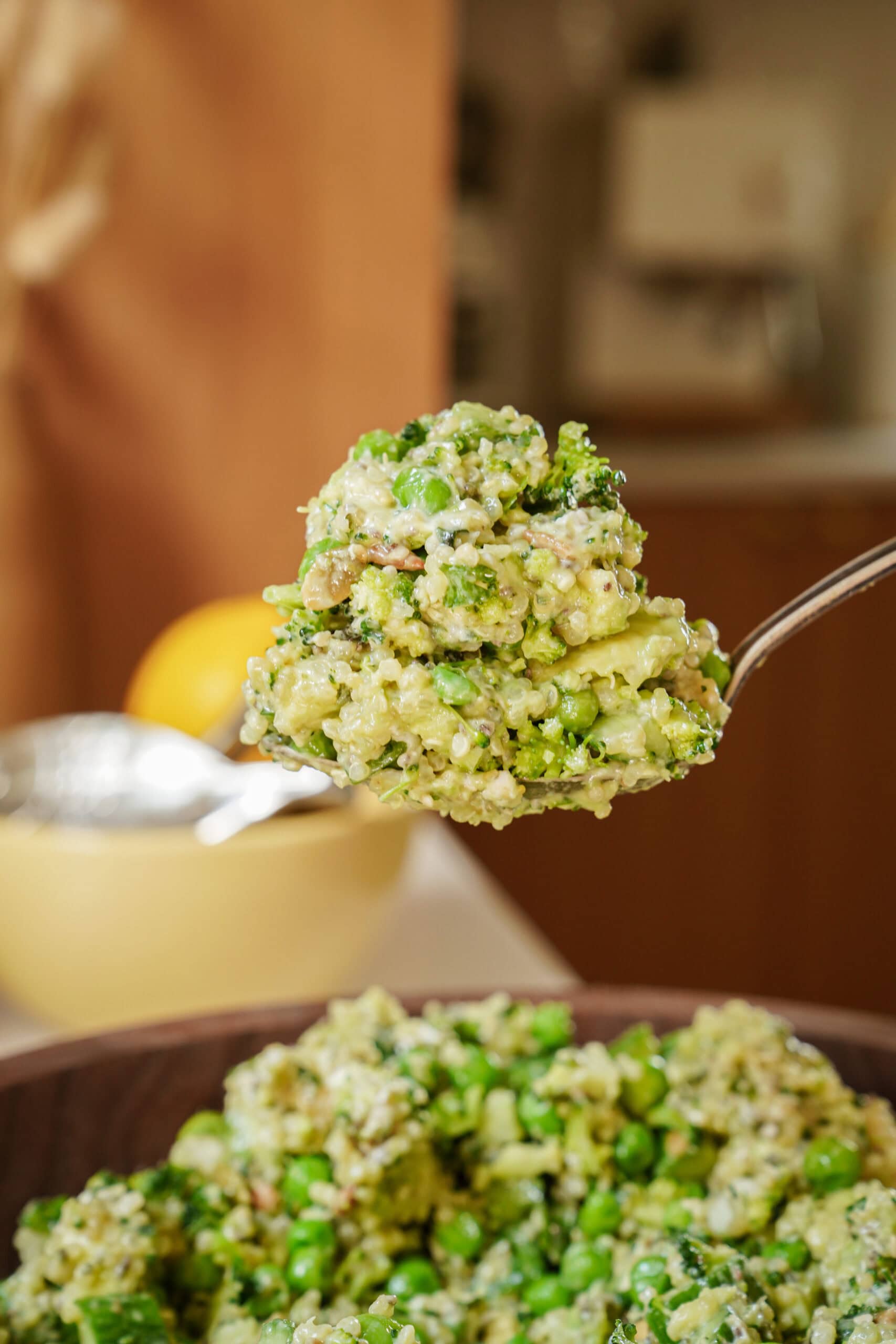 A close-up of a spoonful of quinoa salad with green peas and broccoli, held above a bowl of the same salad. The background is softly blurred, showing a kitchen setting.