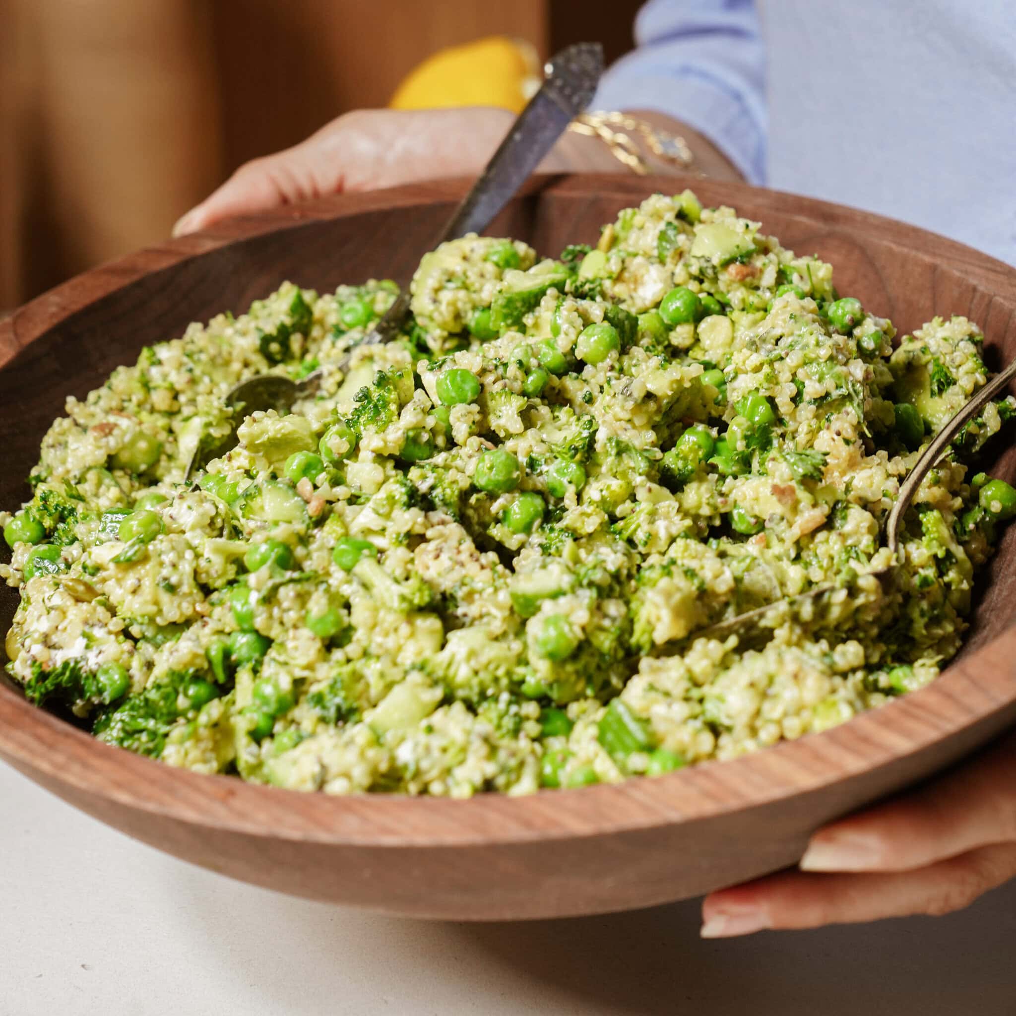 A person holds a wooden bowl filled with a green quinoa salad containing peas, broccoli, and herbs. Two serving spoons are placed in the bowl.