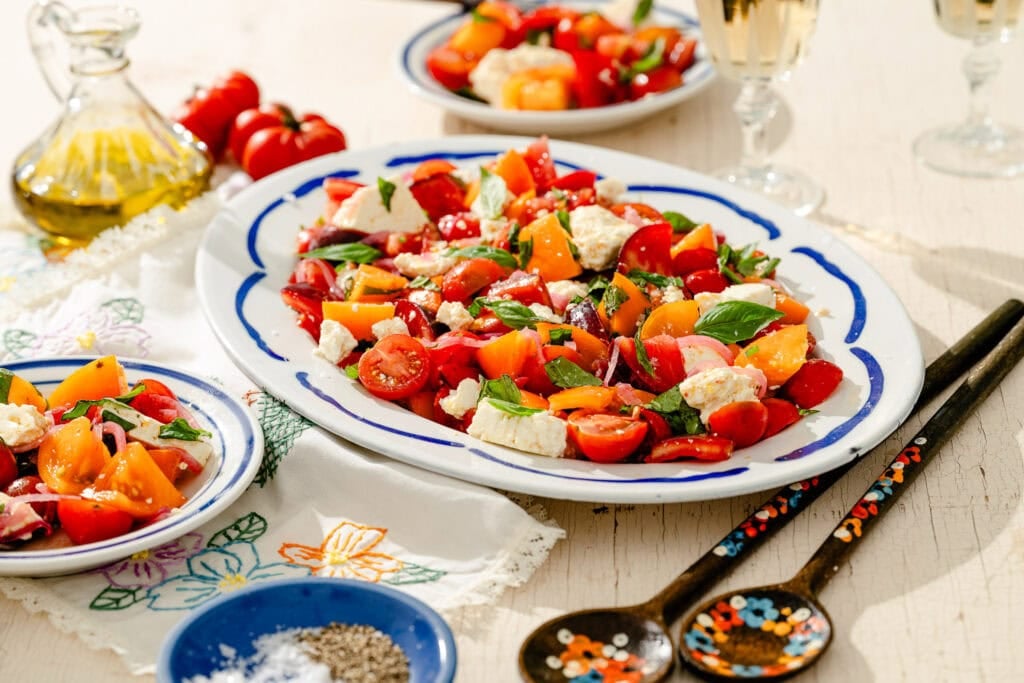 A platter of colorful tomato salad with basil and cheese sits on a table, alongside plates, wooden utensils, a bottle of olive oil, and glasses of sparkling drink.