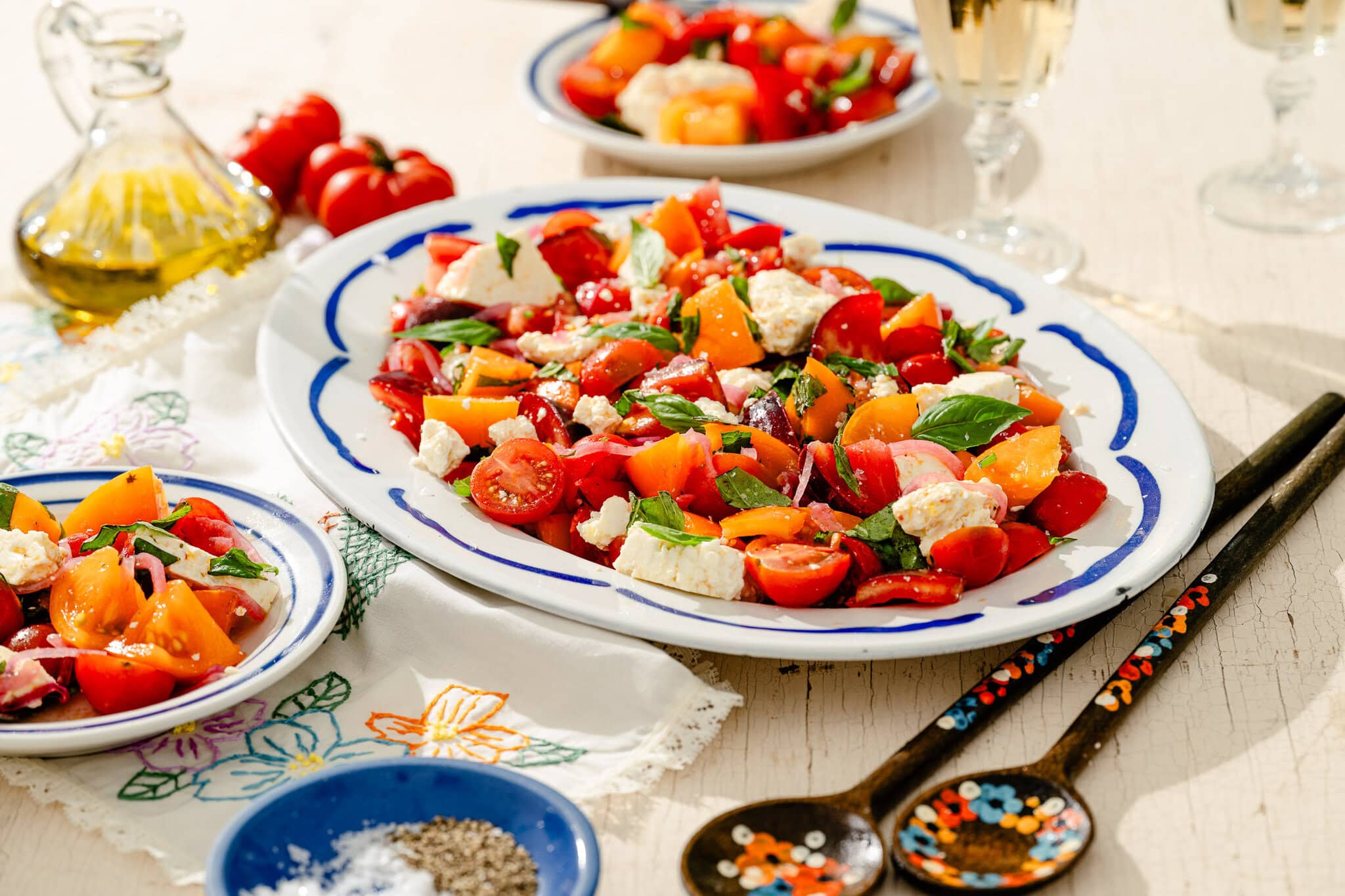 A platter of colorful tomato salad with basil and cheese sits on a table, alongside plates, wooden utensils, a bottle of olive oil, and glasses of sparkling drink.