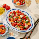 A platter of colorful tomato salad with feta, basil, and herbs sits on a table, surrounded by small plates, glasses of white wine, and wooden salad servers. Bright sunlight highlights the fresh ingredients.