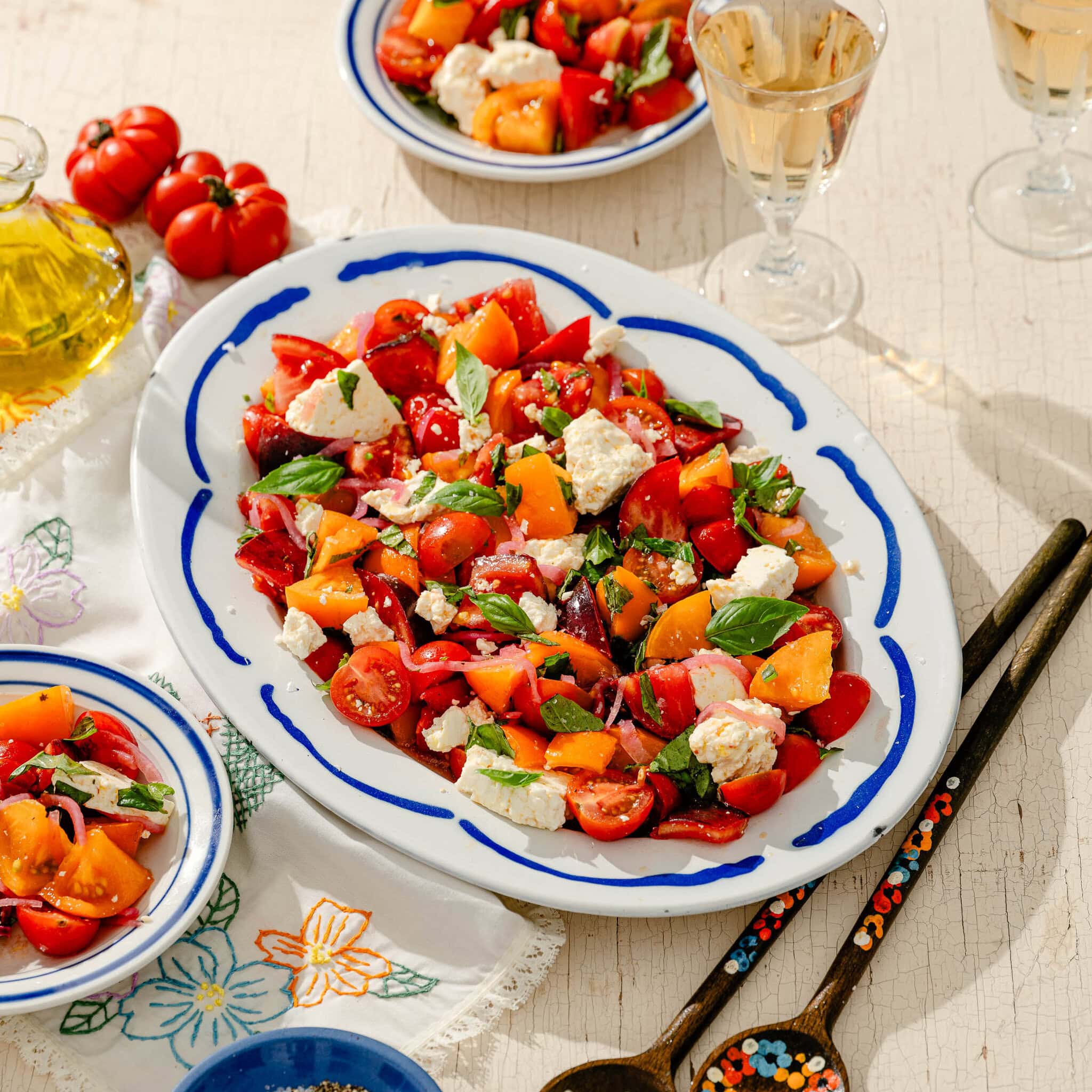 A platter of colorful tomato salad with feta, basil, and herbs sits on a table, surrounded by small plates, glasses of white wine, and wooden salad servers. Bright sunlight highlights the fresh ingredients.