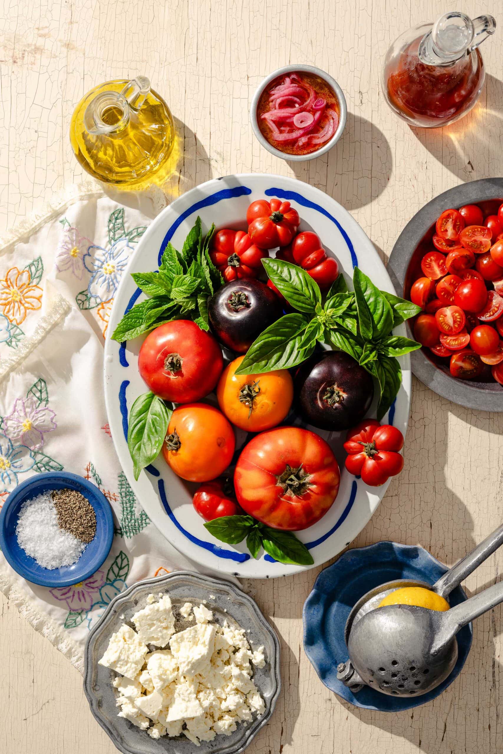 Colorful heirloom tomatoes, basil, and cherry tomatoes on a platter, surrounded by bowls of feta, salt, pepper, pickled onions, oil, and vinegar, with a lemon squeezer and embroidered cloth on a light surface.