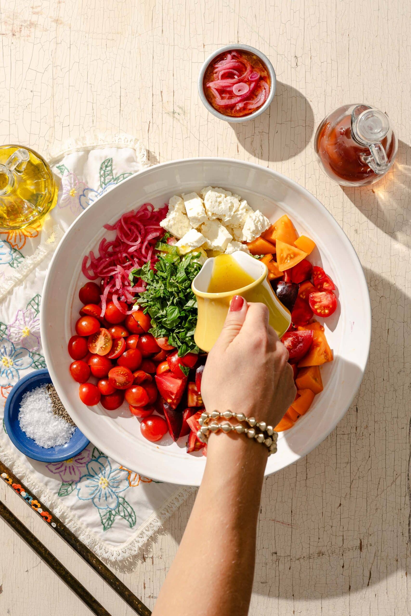 A hand pours dressing over a bowl filled with cherry tomatoes, feta cheese, parsley, red onions, and bell peppers. Other ingredients and a napkin are nearby on a rustic white table.