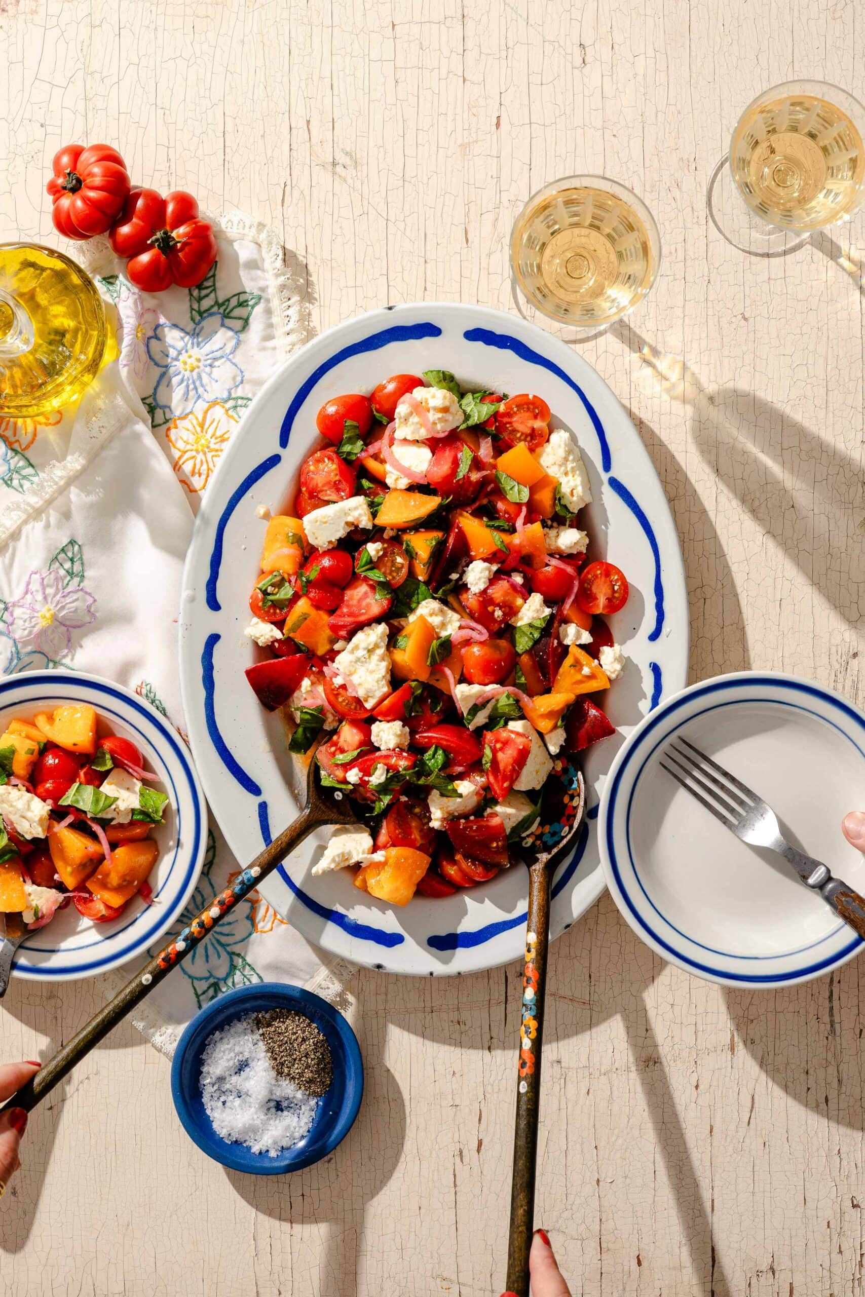 A platter of colorful tomato salad with feta cheese and basil, surrounded by two bowls, a small dish of pepper, glasses of white wine, and a floral napkin on a rustic table.