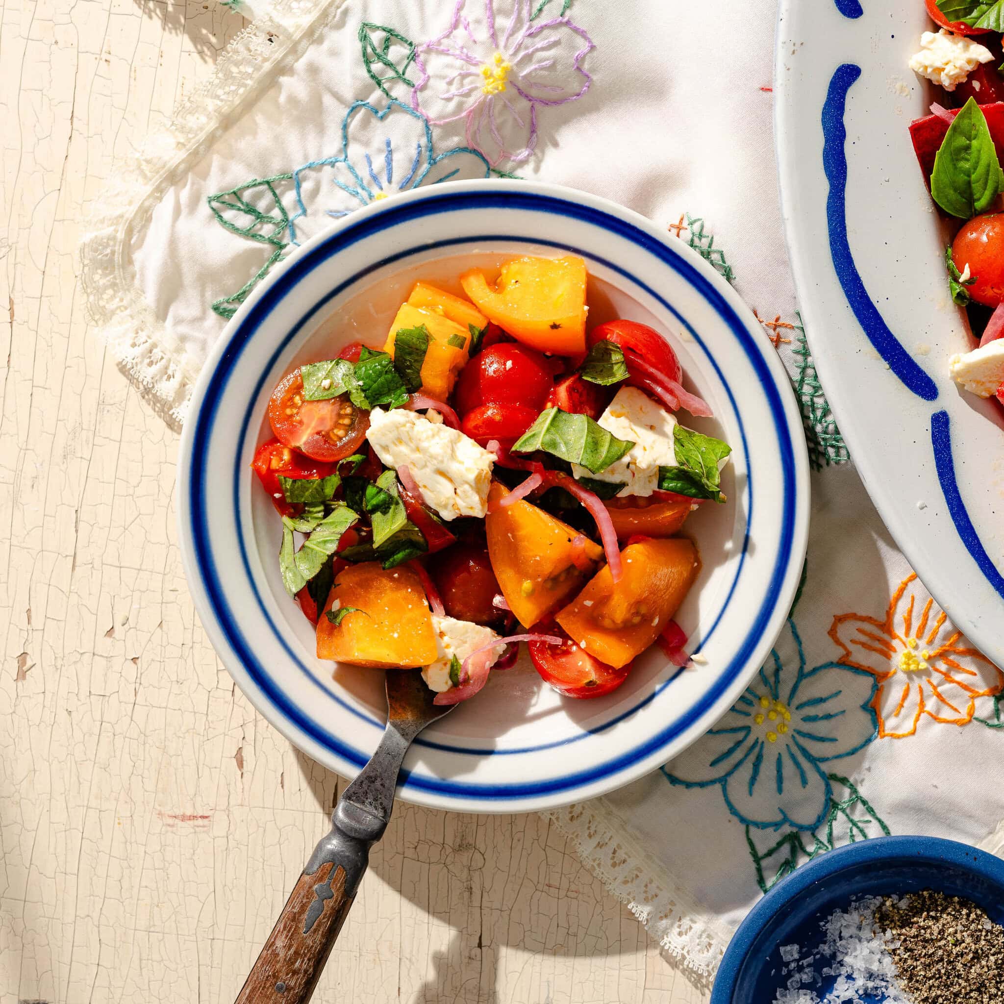 A bowl of salad with yellow and red tomatoes, feta cheese, onions, and fresh basil on a white plate with blue stripes. The bowl rests on a floral-embroidered cloth. A fork is placed inside the bowl.