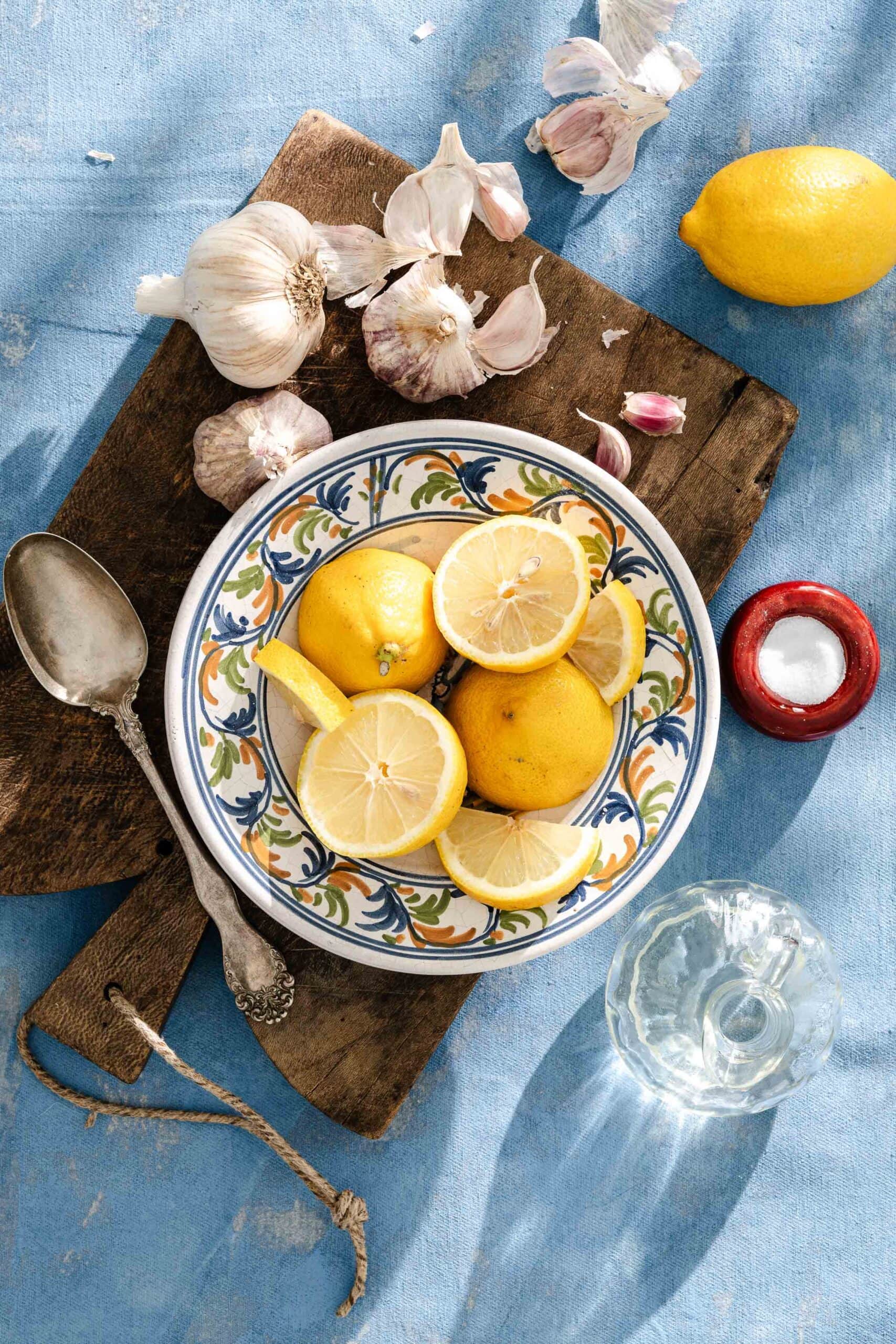 A decorative bowl with whole and sliced lemons sits on a wooden board, surrounded by garlic, a spoon, a glass of water, a small salt dish, and a lemon, all on a blue textured surface.