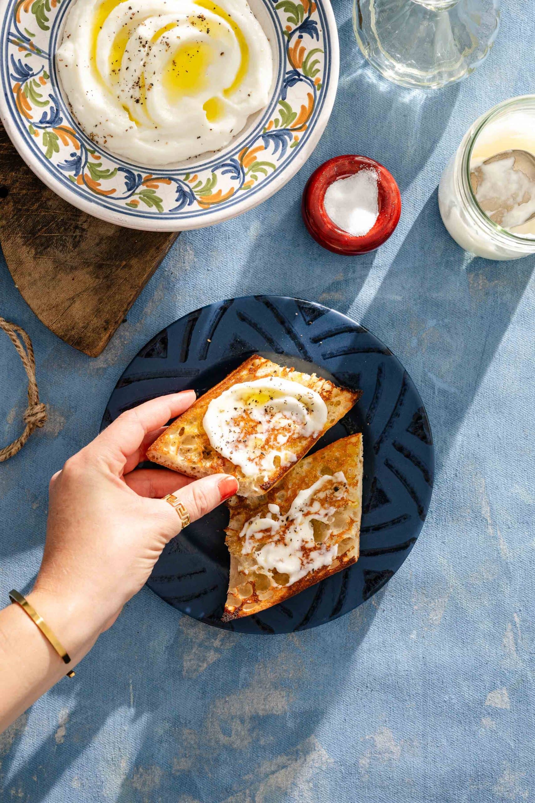 A hand holding toast spread with labneh, on a blue plate. A bowl of labneh with olive oil, salt jar, and water glass are nearby on a textured blue surface in sunlight.