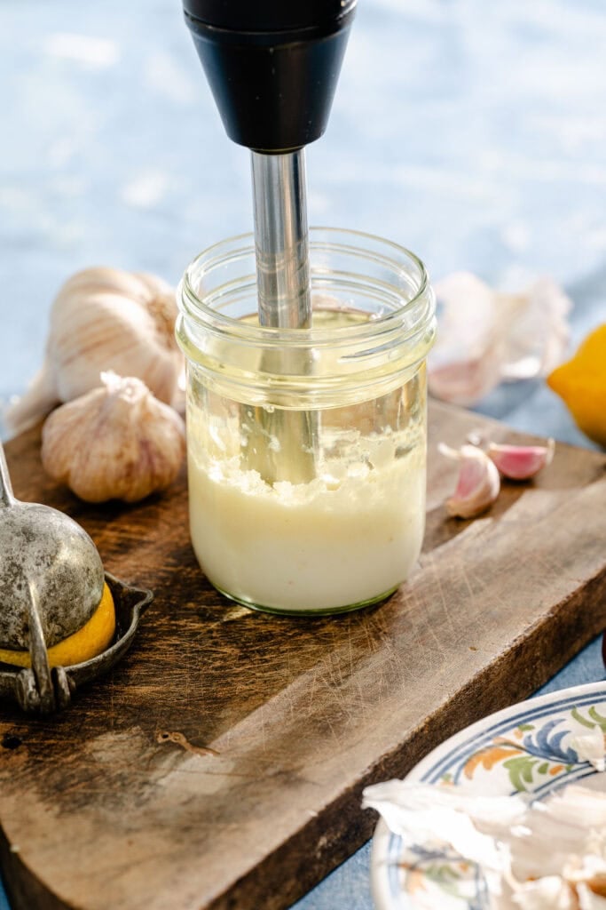 A hand blender mixes a creamy mixture in a glass jar on a wooden board, surrounded by garlic, a lemon, a garlic press, and a decorative plate.
