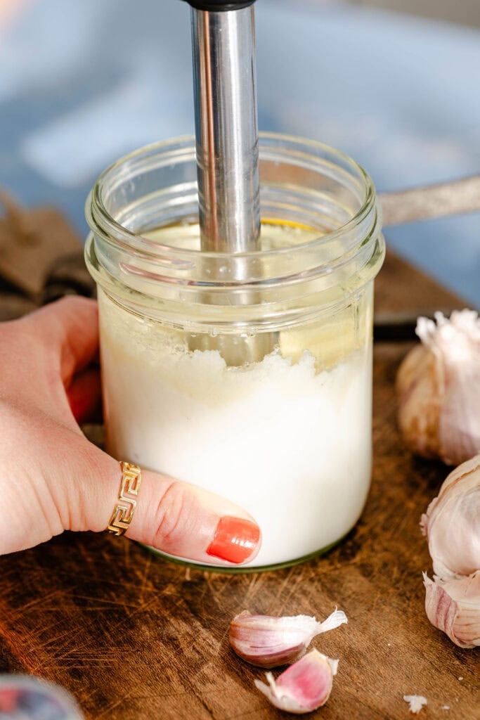 A hand holds a glass jar with white creamy mixture being blended using an immersion blender; garlic bulbs and cloves sit on a wooden surface nearby.