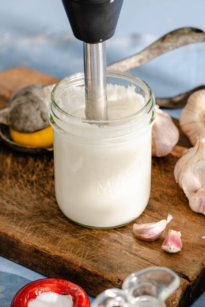 A hand blender blending a creamy white mixture in a mason jar on a wooden board, surrounded by garlic cloves, a lemon squeezer, and kitchen items.