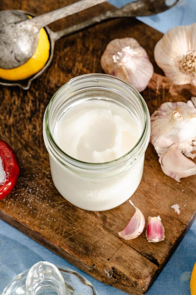 A jar of creamy white spread on a wooden board, surrounded by garlic bulbs, cloves, a lemon, and a metal citrus squeezer.