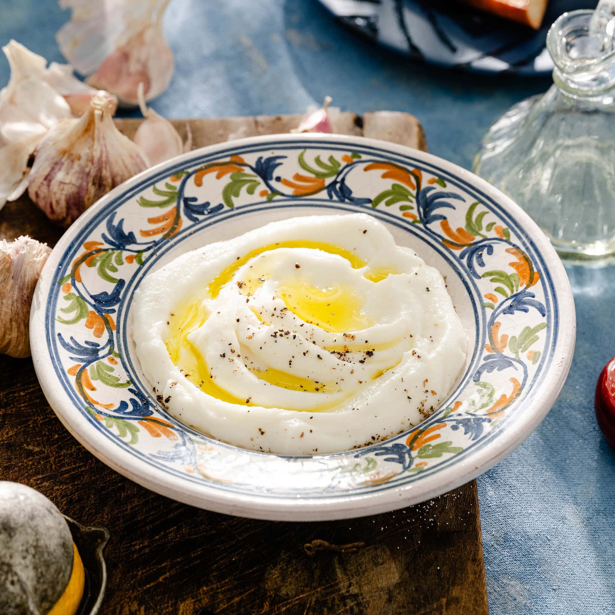 A colorful plate with creamy white mashed potatoes drizzled with olive oil and sprinkled with black pepper, placed on a wooden surface with garlic cloves nearby.