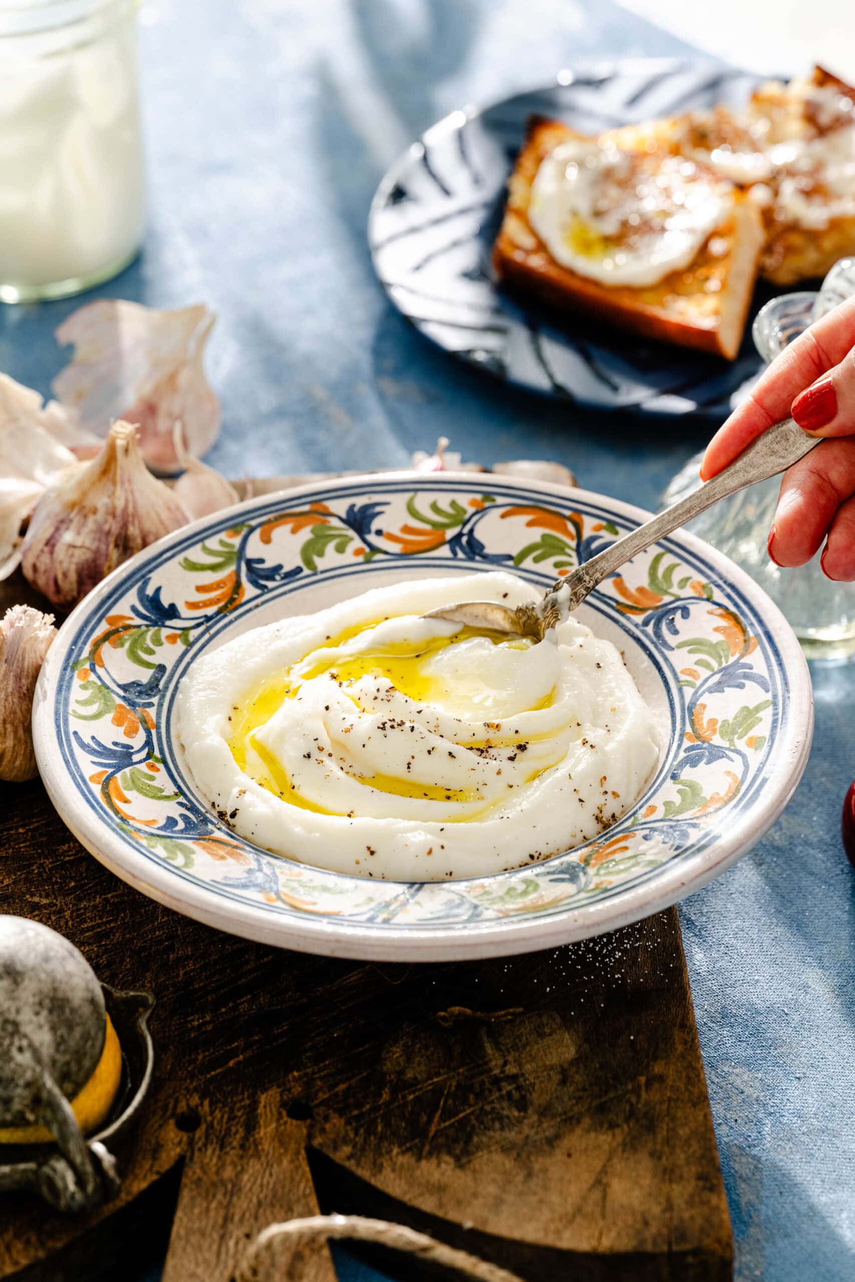 A hand spoons creamy yogurt in a patterned bowl, drizzled with olive oil and pepper. Garlic, lemon, and toast with an egg are nearby on a rustic wooden board and blue tablecloth.