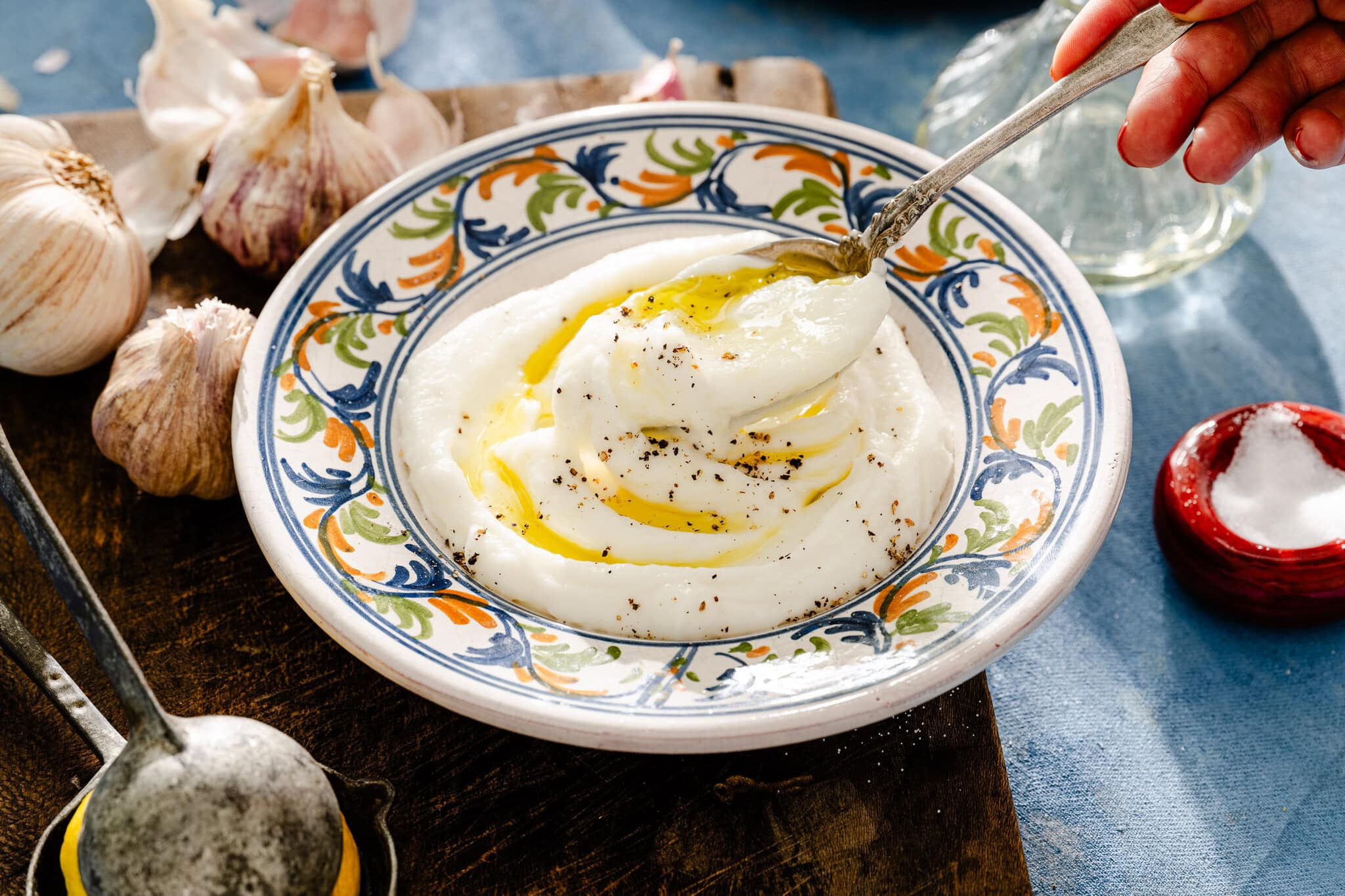 A colorful bowl of creamy dip topped with olive oil and pepper sits on a wooden board with garlic cloves nearby; a hand holds a spoon over the dip.