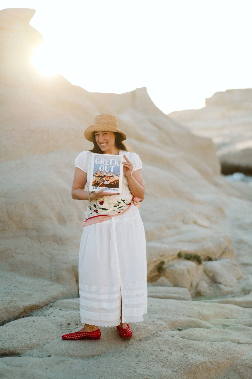 A woman in a white dress, red shoes, and a straw hat stands on rocky terrain, smiling and holding a book titled Greek Out with sunlight shining behind her.