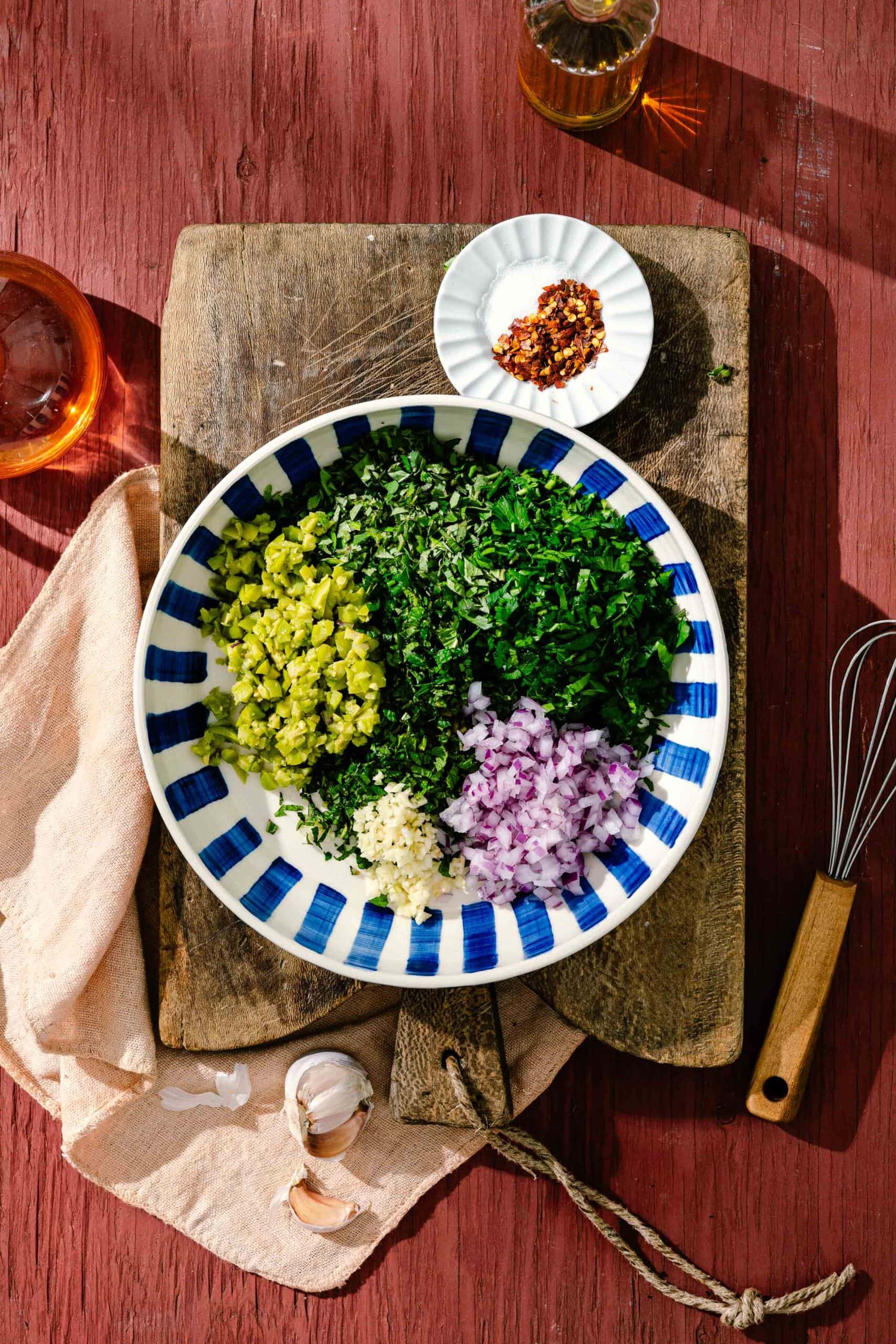 A bowl with chopped parsley, oregano, pickles, red onion, and garlic sits on a wooden board. Nearby are a whisk, garlic cloves, a napkin, a small dish of spices, and glasses of amber liquid on a red wooden table.