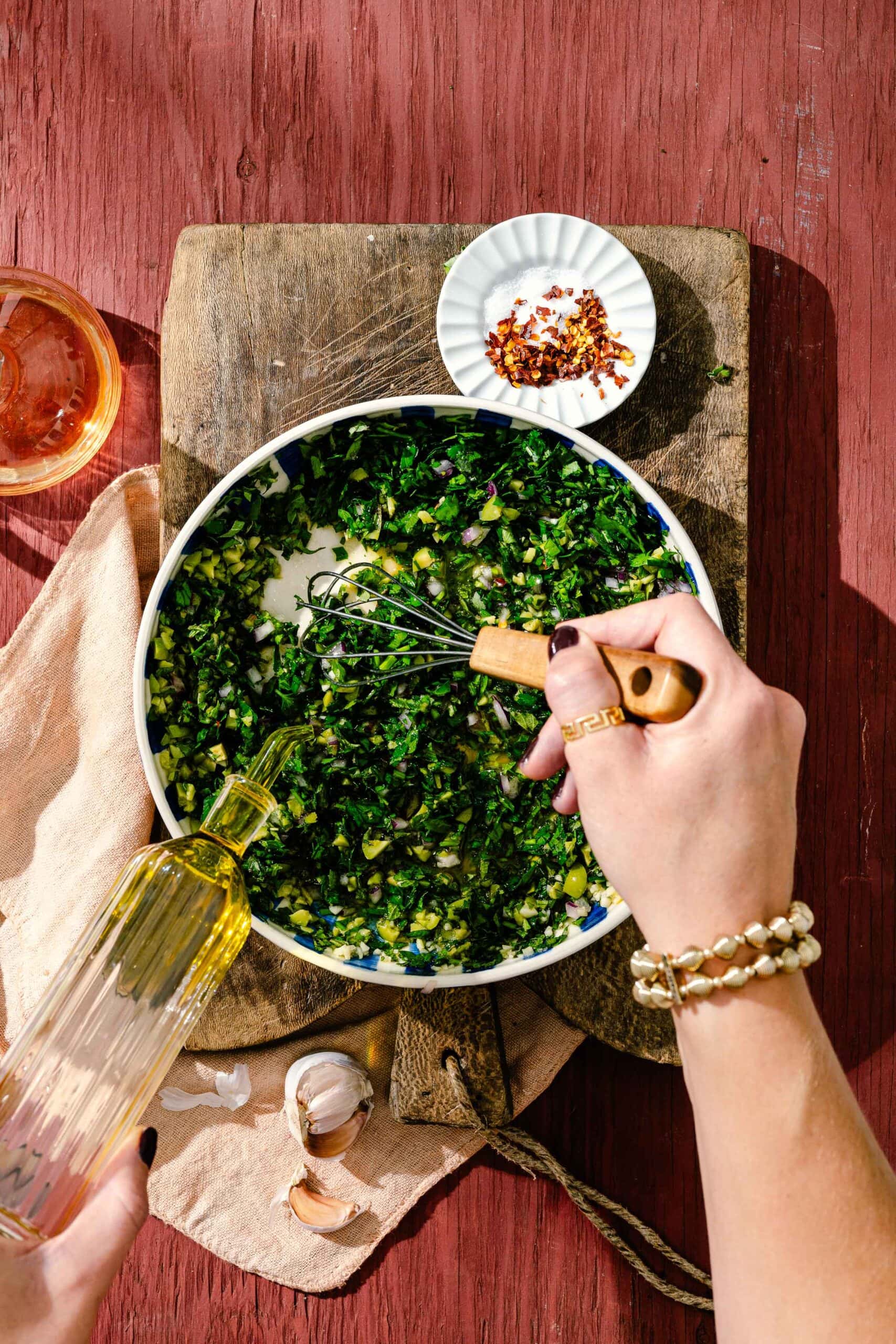 A person whisks chopped greens in a bowl on a wooden board, pouring oil in, with garlic cloves, red pepper flakes, and a drink nearby on a rustic red table.