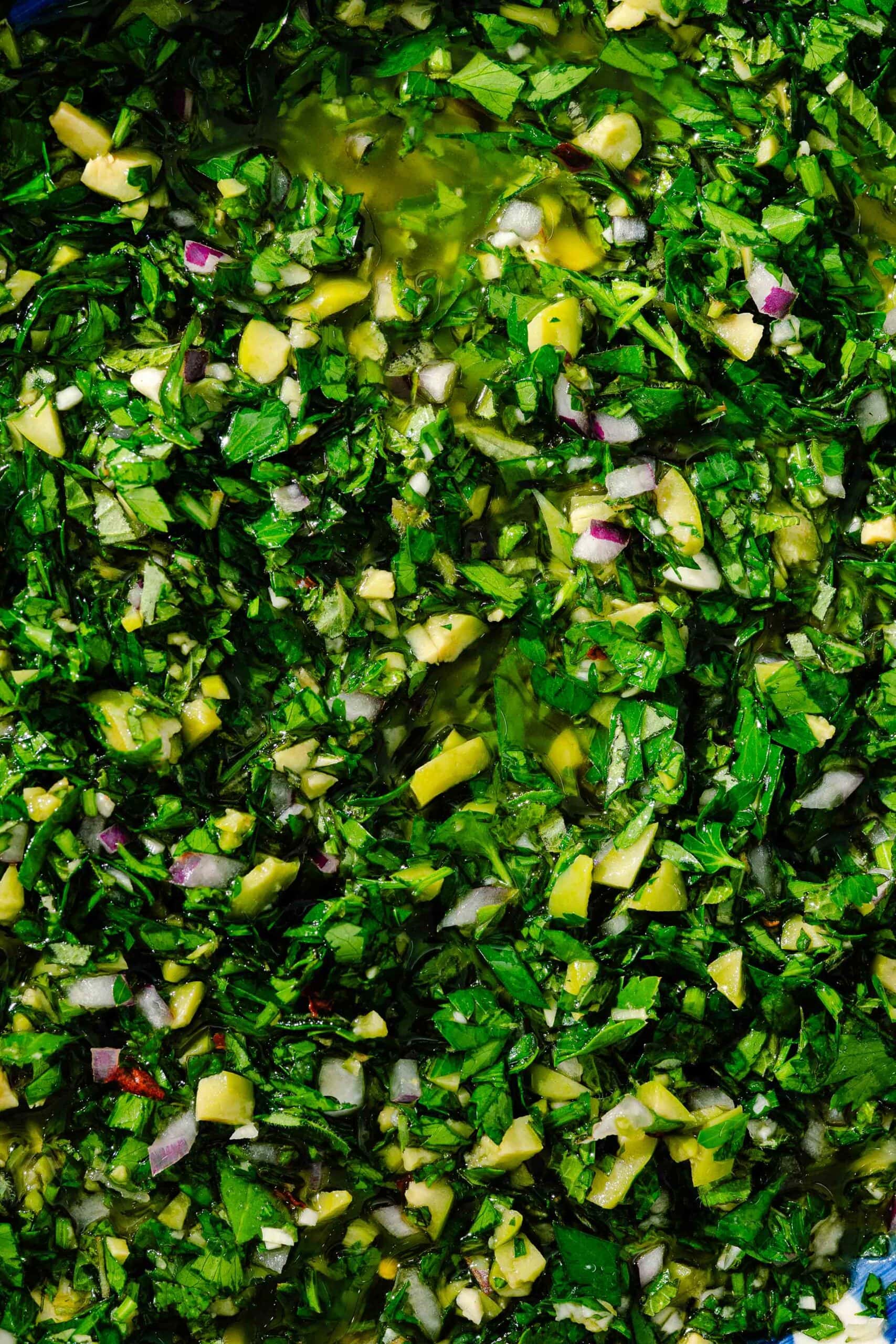 Close-up of chopped fresh herbs, garlic, red onion, and olive oil mixed together, possibly a chimichurri or herb salsa, with a vibrant green and yellow color.