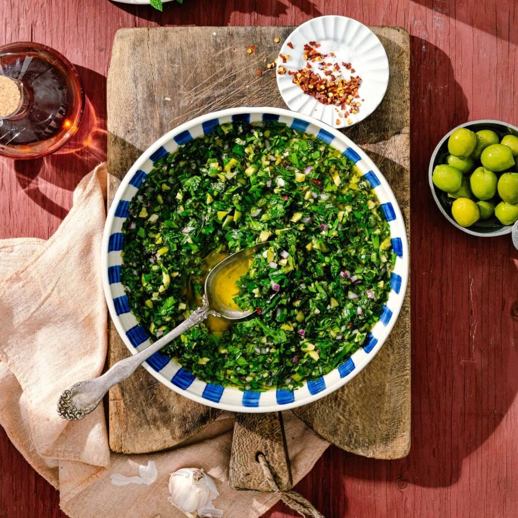 A bowl of green chimichurri sauce with a spoon, set on a wooden board. Nearby are green olives, crushed red pepper on a small plate, a garlic clove, a beige napkin, and a glass bottle with red liquid.