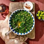 A bowl of green chimichurri sauce with a spoon, set on a wooden board. Nearby are green olives, crushed red pepper on a small plate, a garlic clove, a beige napkin, and a glass bottle with red liquid.