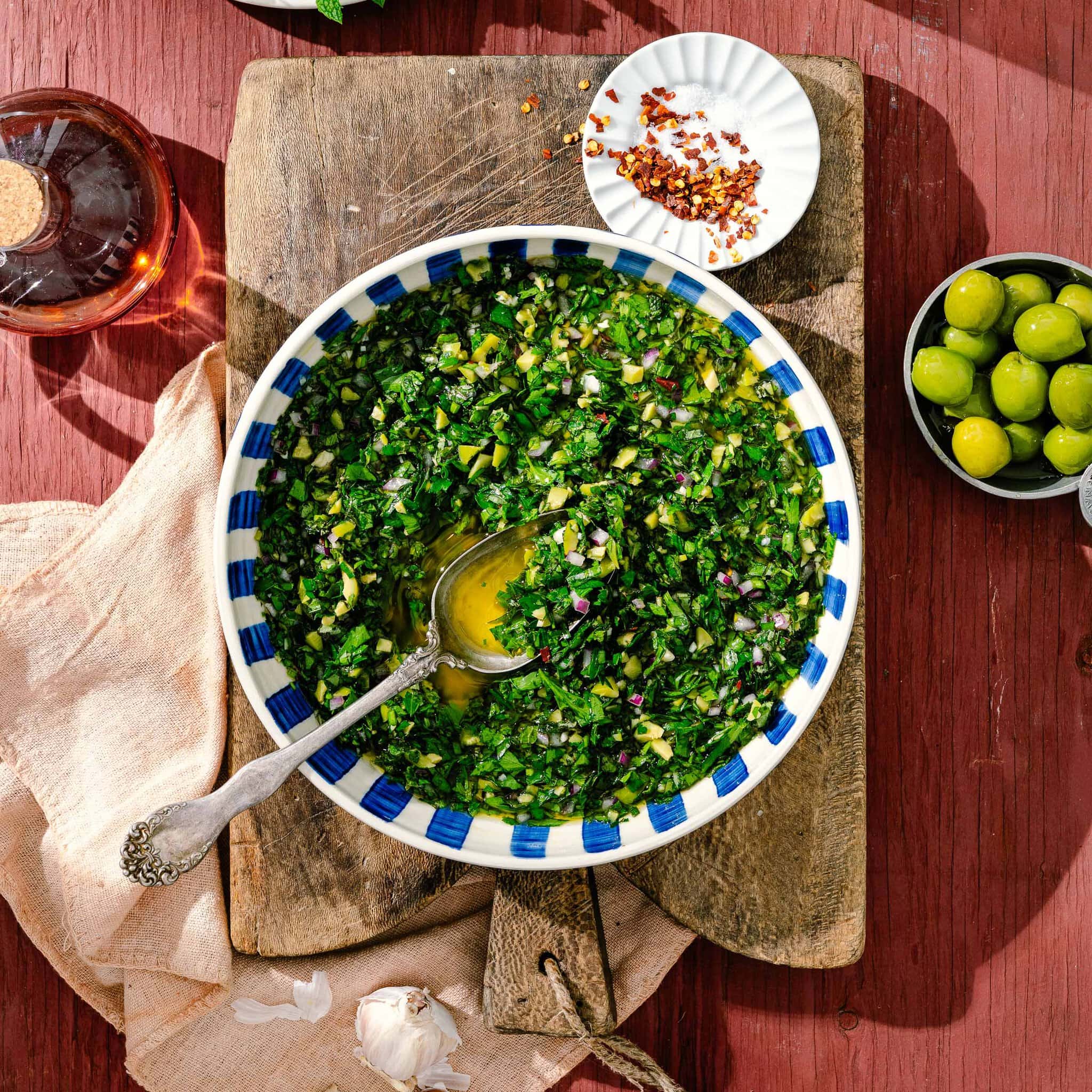 A bowl of green chimichurri sauce with a spoon, set on a wooden board. Nearby are green olives, crushed red pepper on a small plate, a garlic clove, a beige napkin, and a glass bottle with red liquid.