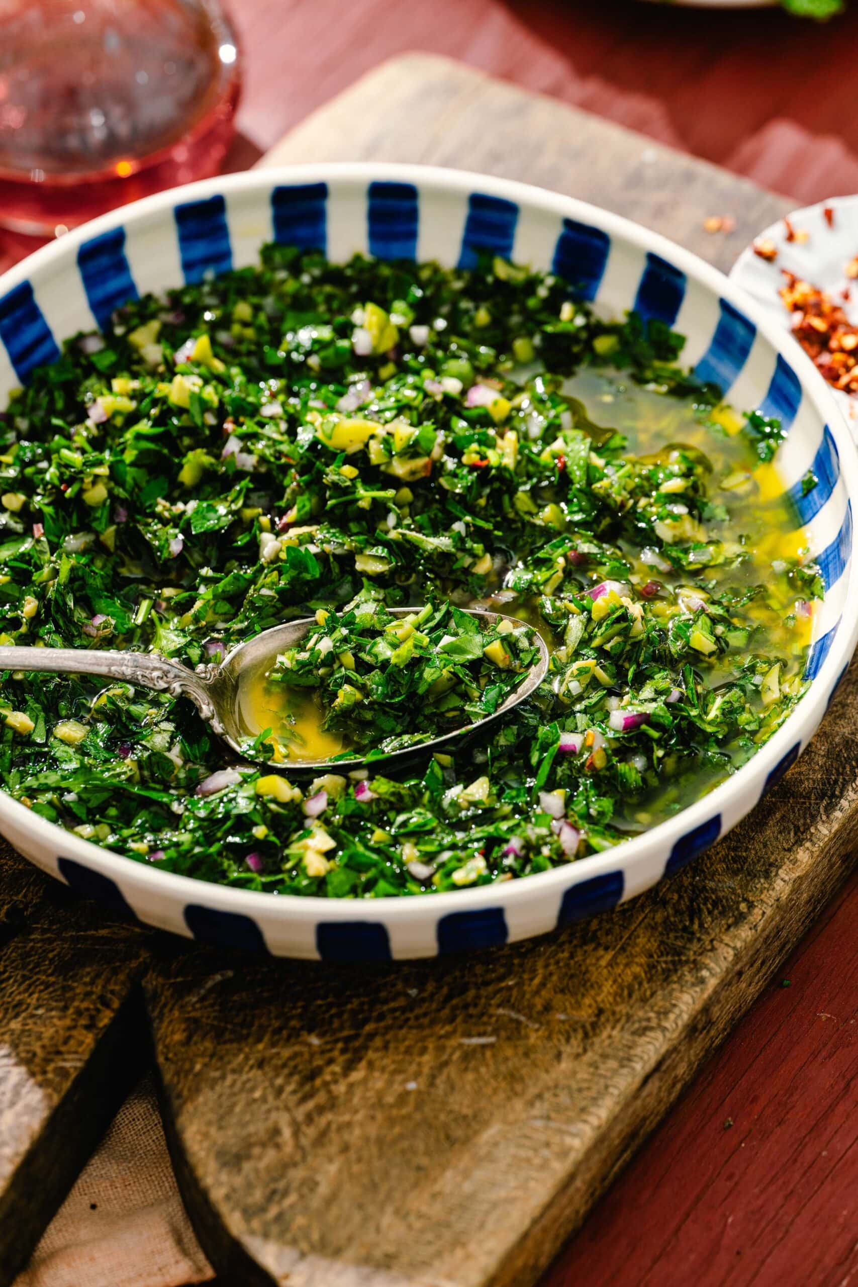 A bowl of chimichurri sauce with chopped herbs, red onion, and oil sits on a wooden board. A spoon rests in the bowl. The bowl has a blue and white pattern.