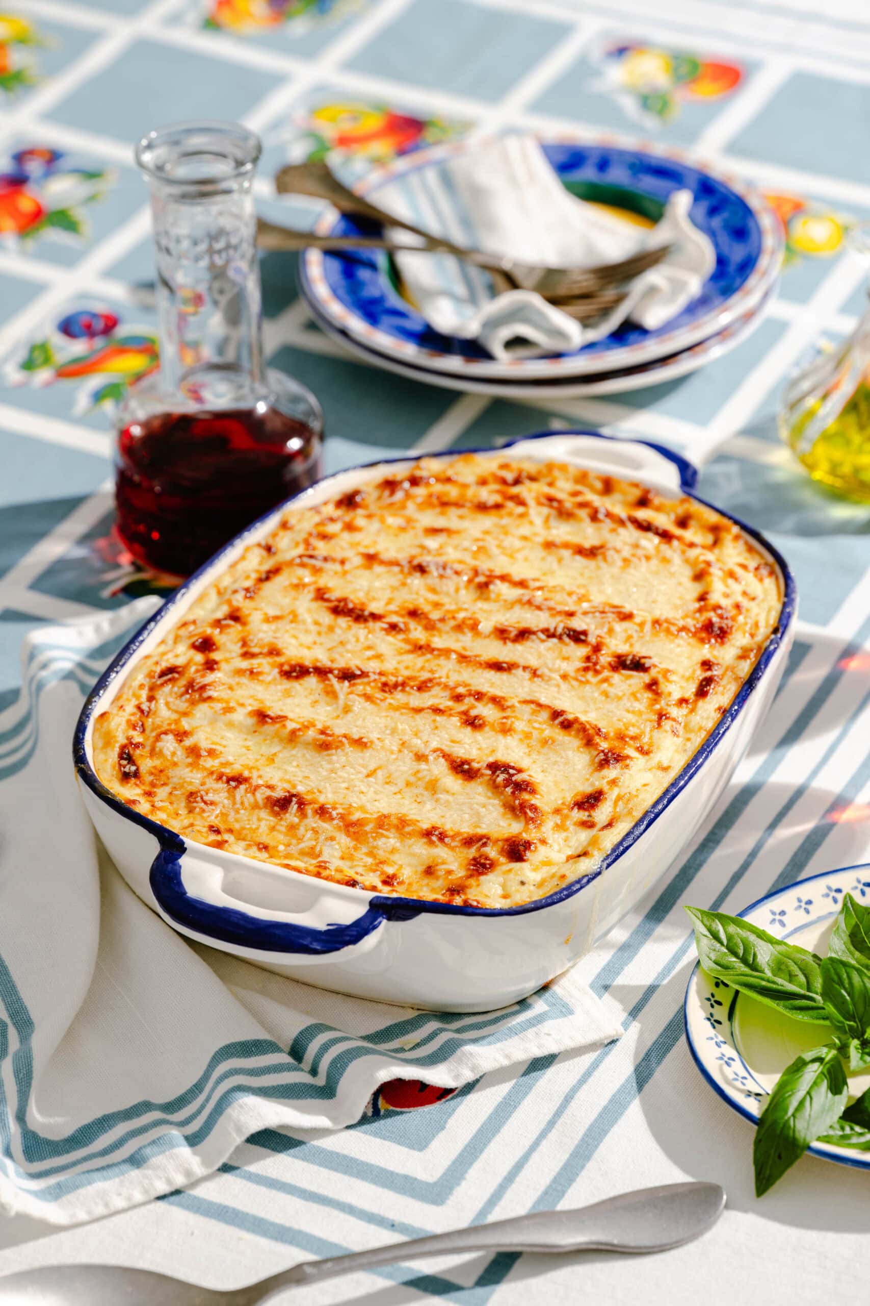 A white ceramic baking dish filled with a golden, cheese-topped casserole sits on a table with a patterned blue and white tablecloth. Nearby are a small carafe of red wine, a plate with fresh basil, and stacked plates with silverware.