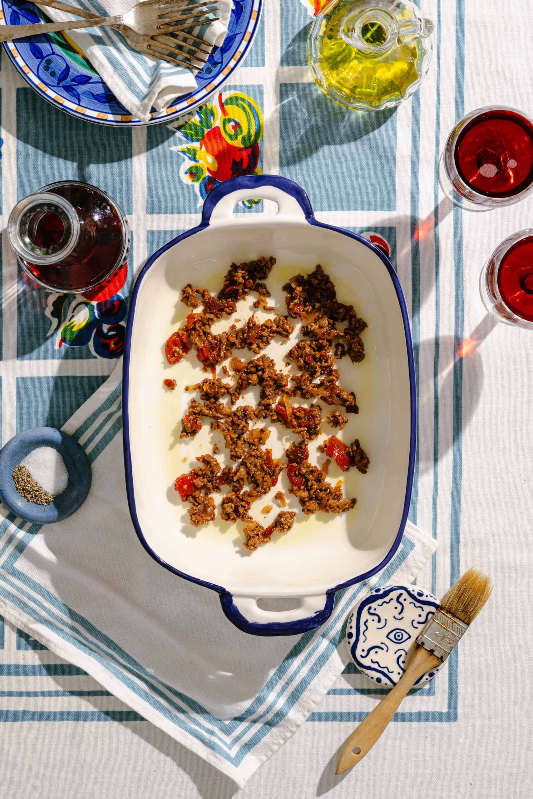 A baking dish with ground meat and tomato topping sits on a patterned tablecloth, surrounded by wine glasses, oils, pepper, a brush, utensils, and a plate. Sunlight casts shadows across the scene.